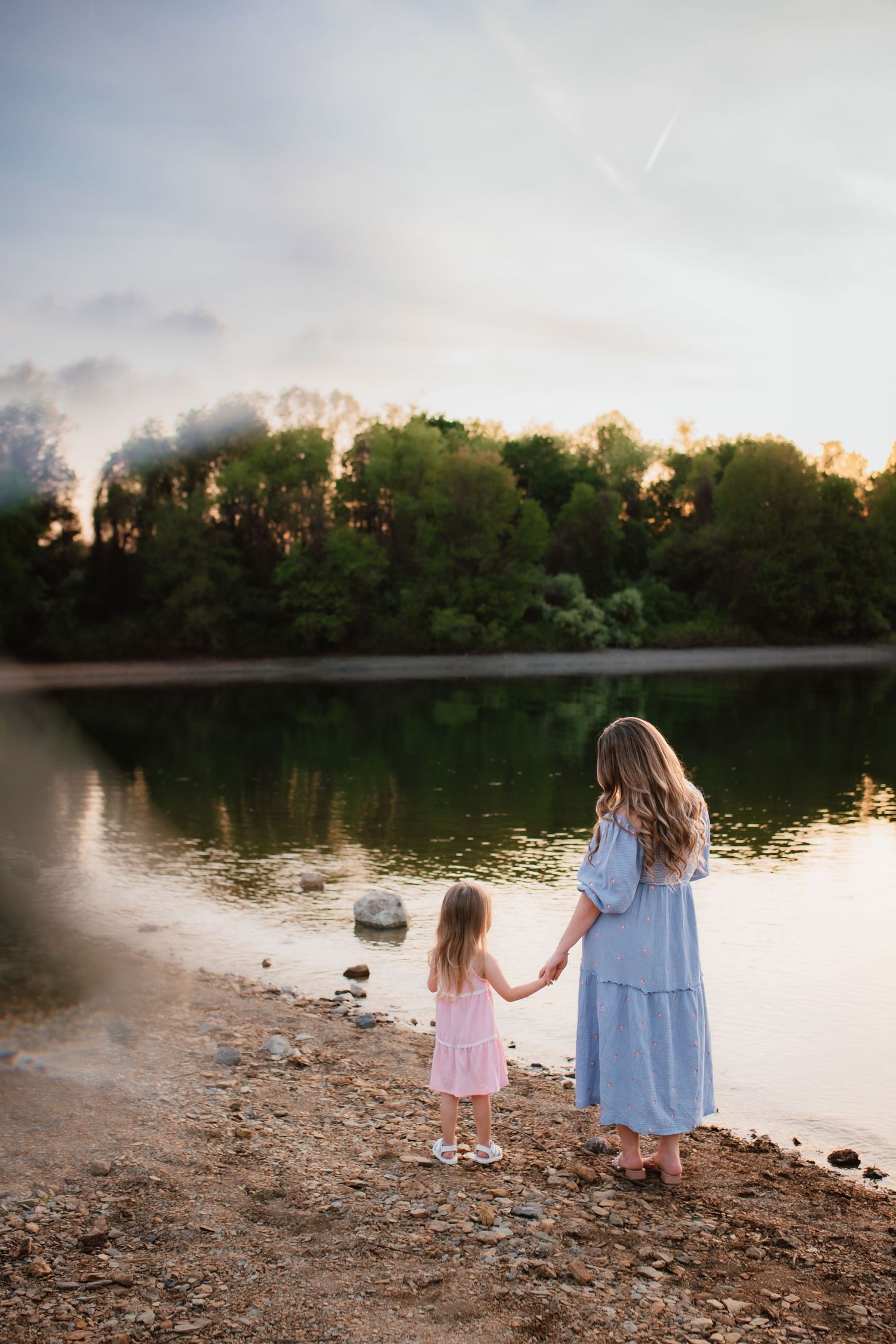 Mother and young daughter holding hands while gazing at tranquil lake at sunset.