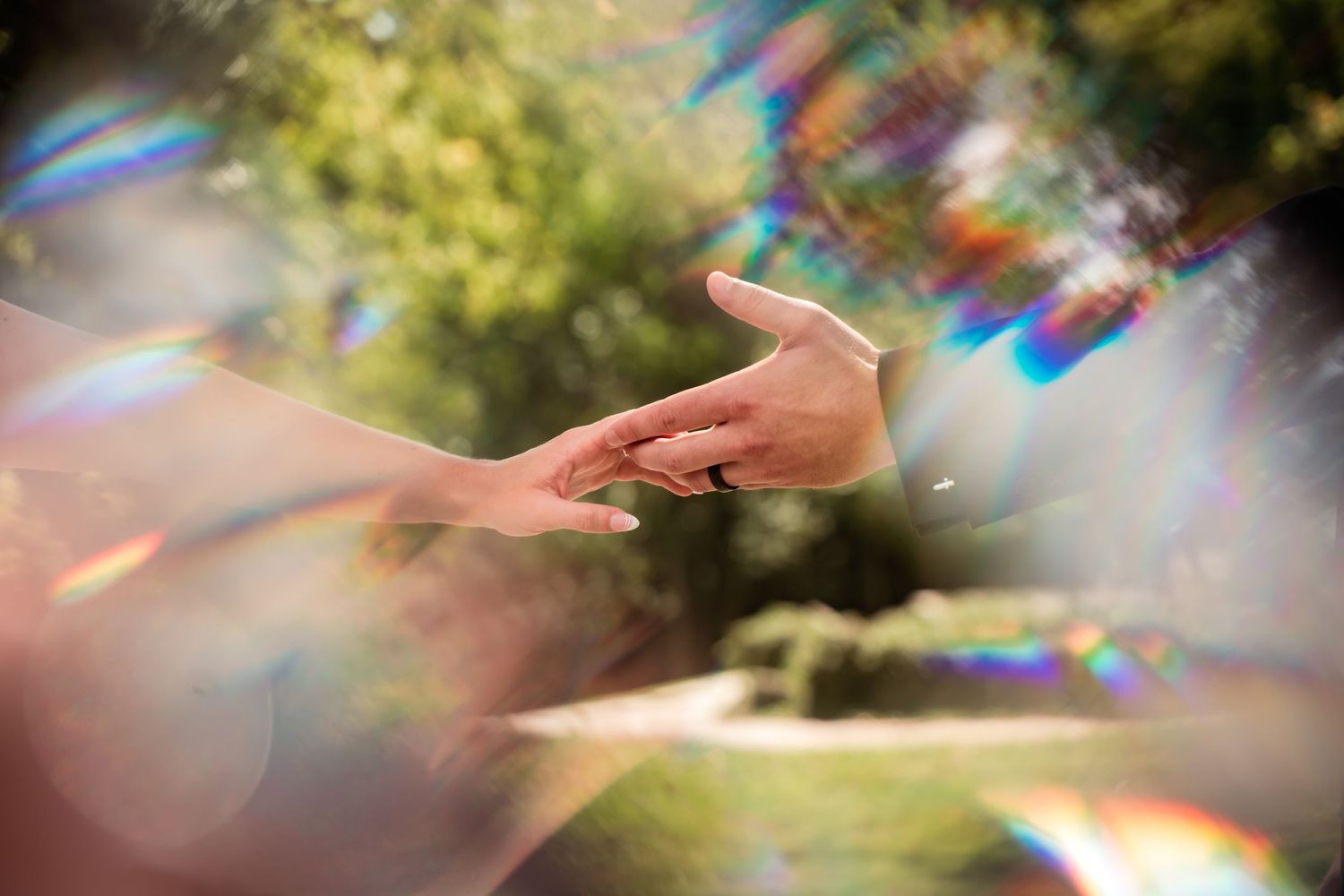 Two hands reaching toward each other surrounded by colorful lens flare bokeh in a sunlit garden.
