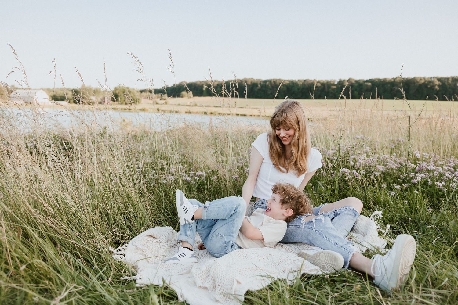 Mother and child relaxing on blanket in tall grass meadow near lake on sunny day.