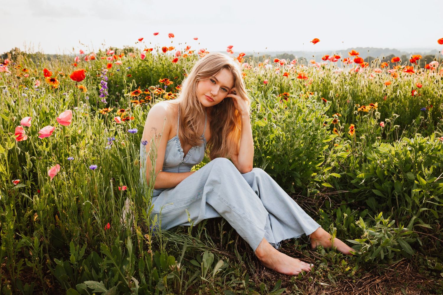 Beautiful blonde woman sitting barefoot in a vibrant poppy flower field wearing blue overalls.