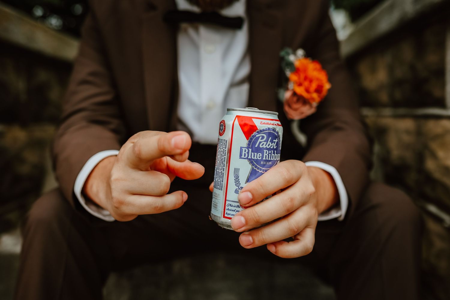 Groom in brown suit holding a Pabst Blue Ribbon beer can toward camera.