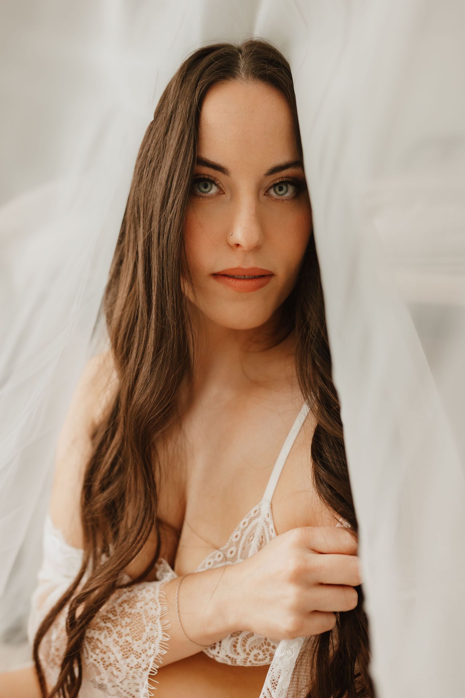 Brunette woman with long hair in white lace lingerie gazing through sheer white fabric curtain.