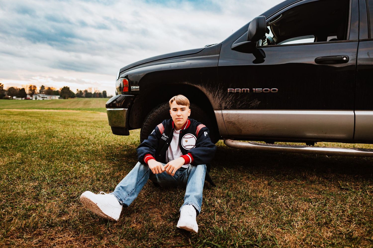 Teen boy in varsity jacket sits against black Ram 1500 truck on green field for senior portrait.