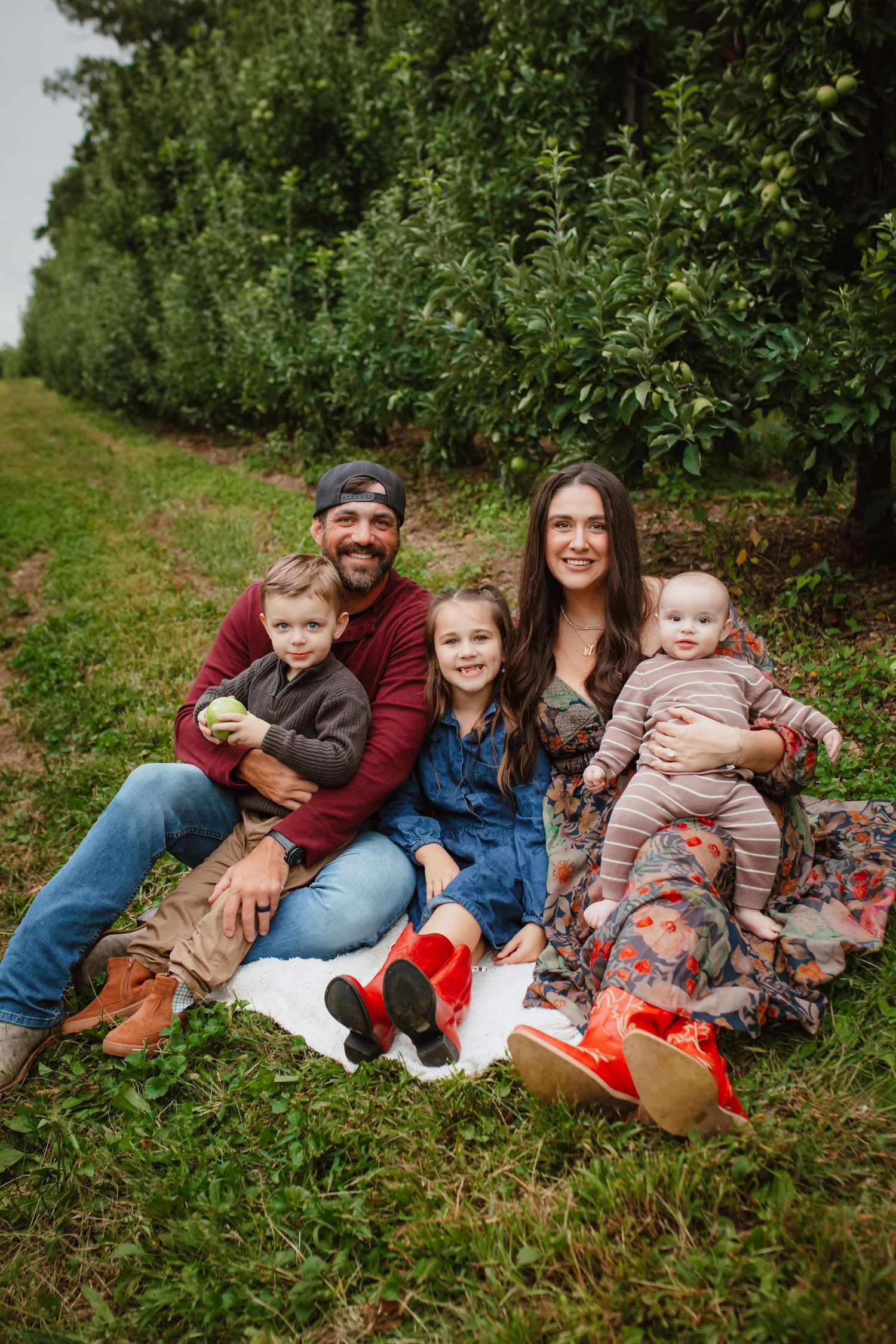 Smiling family of five sits on a white blanket in a grassy apple orchard, with two parents, a toddler, a girl in red boots, and a baby.