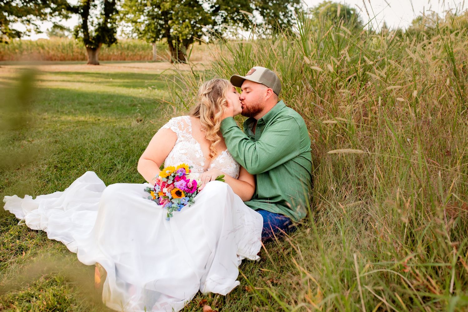 Bride and groom share a romantic kiss while sitting in tall grass, bride holding a colorful wildflower bouquet.