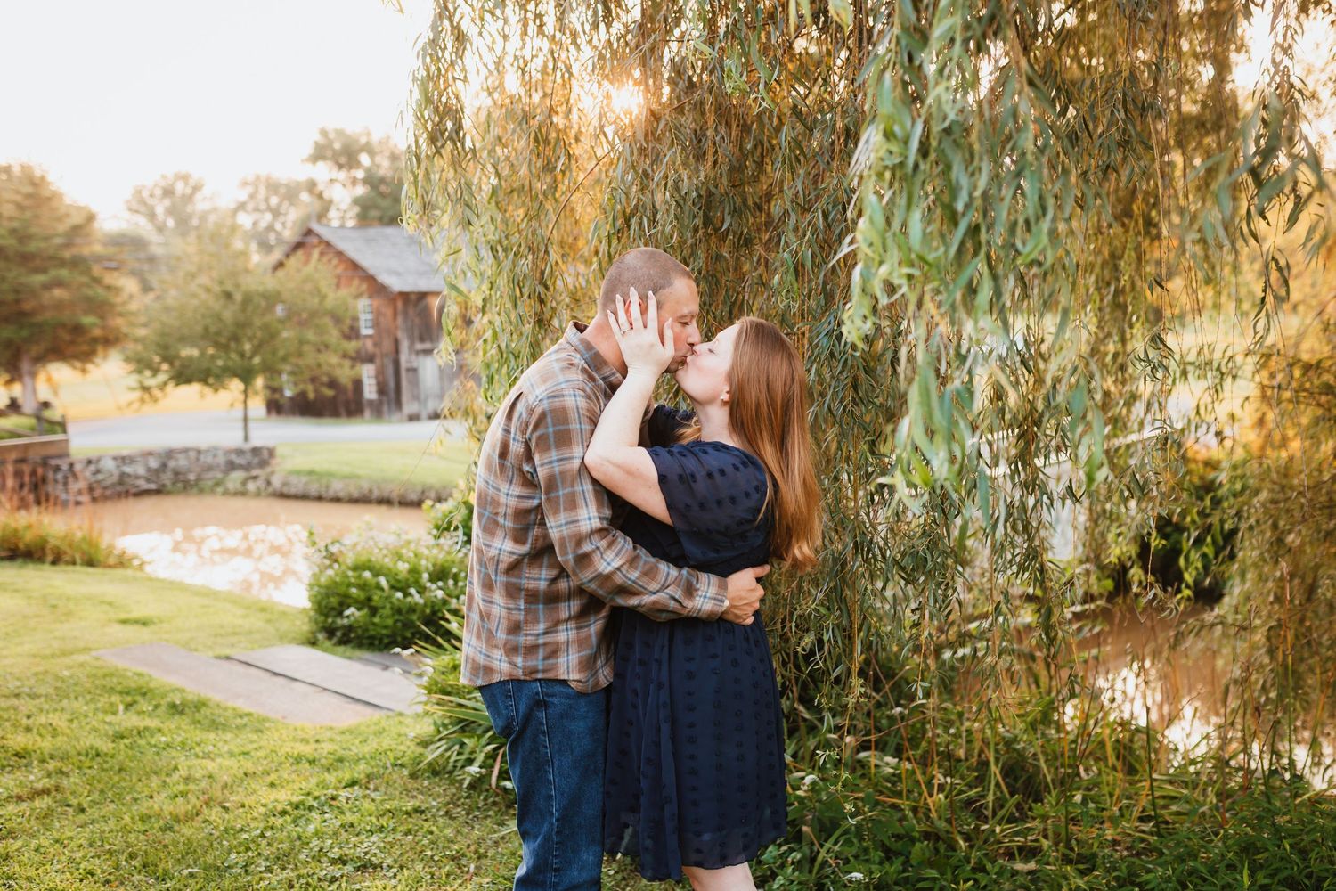 Couple sharing a kiss under a weeping willow tree beside a pond at golden hour in a scenic garden.