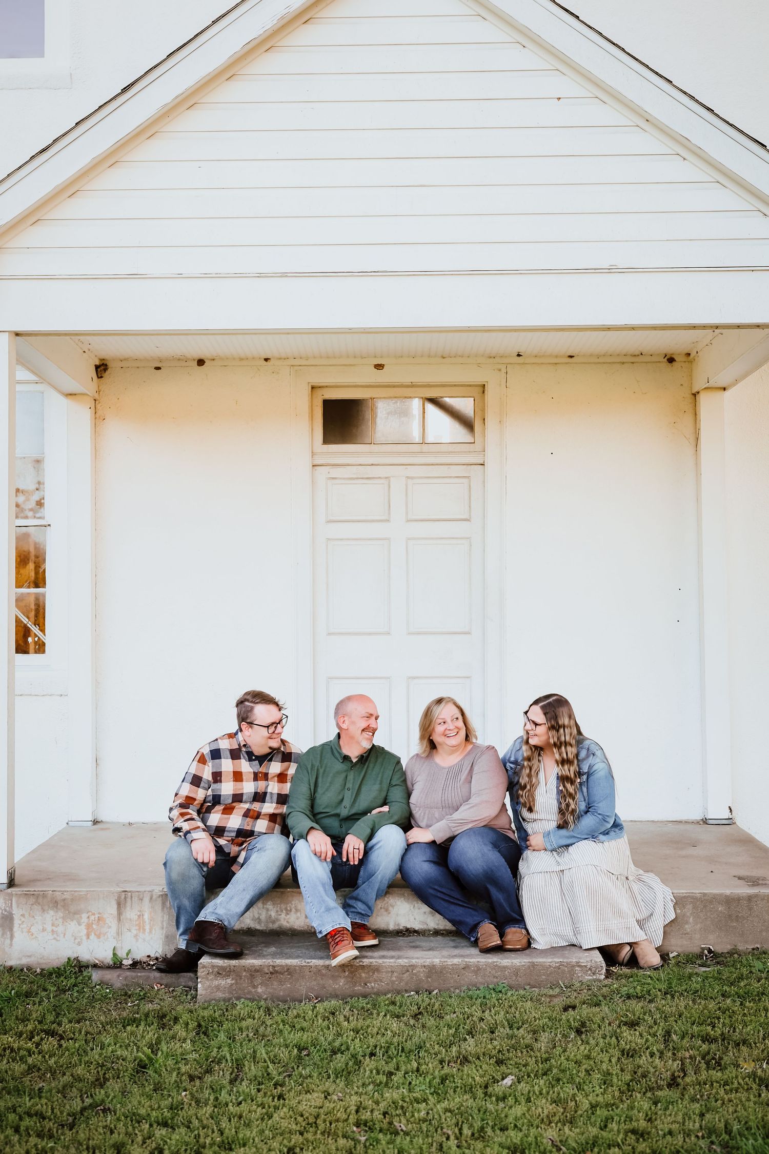 Family of four sits casually on white church steps smiling and laughing together.