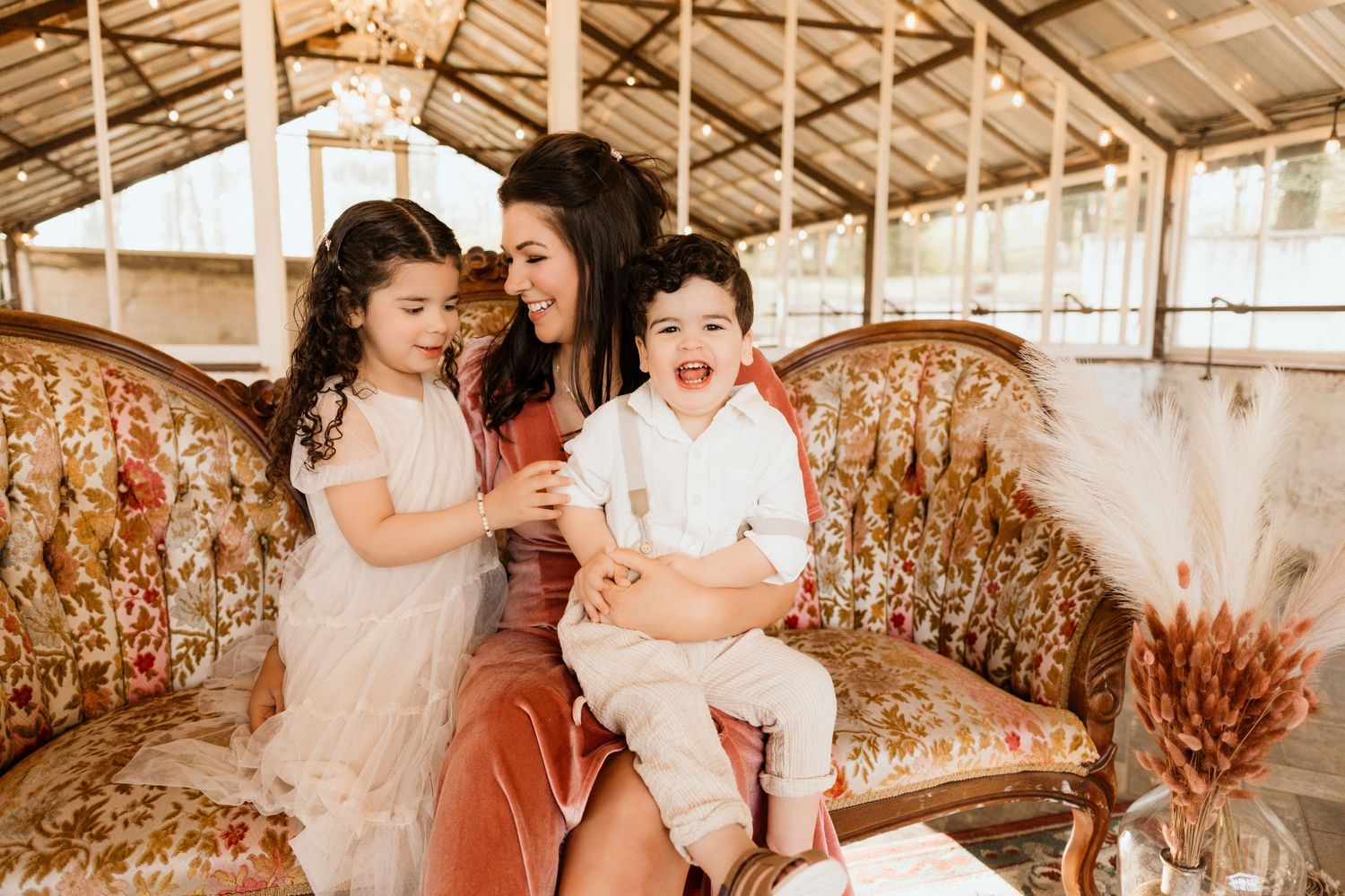A mother in a pink dress laughs with her two young children on a vintage sofa.