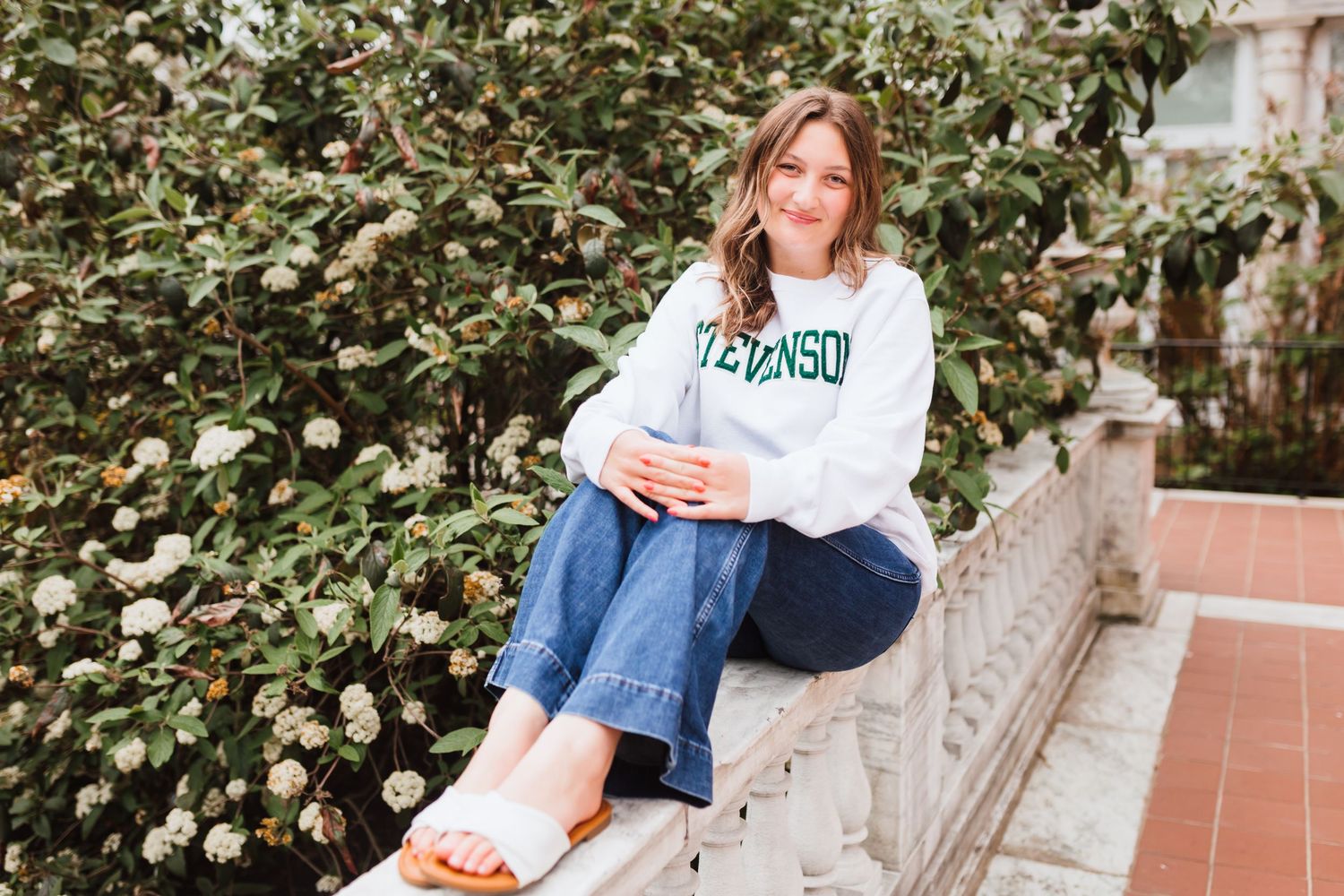 Young woman in Stevenson sweatshirt and jeans sits on stone ledge surrounded by white flowering bushes.