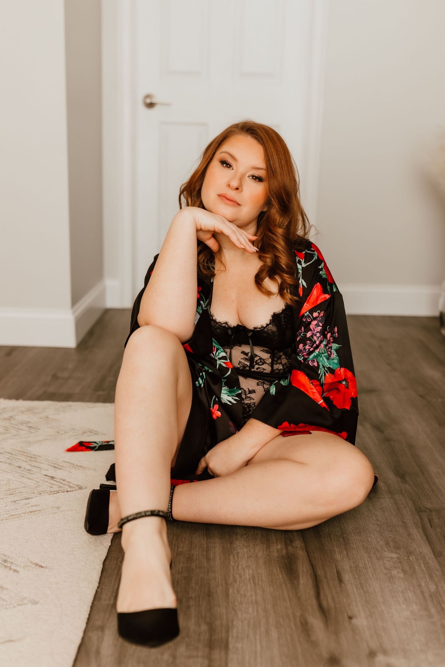 Redhead woman in floral kimono and black lace lingerie sits on hardwood floor.