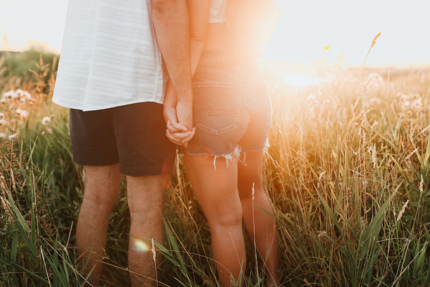 Couple holding hands in golden sunlit tall grass field during outdoor engagement photo session.