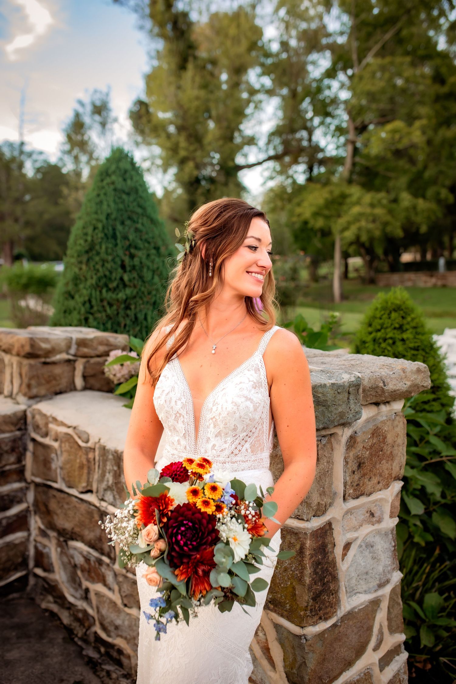 Smiling bride in lace gown holds a vibrant red and white bouquet beside a stone wall.