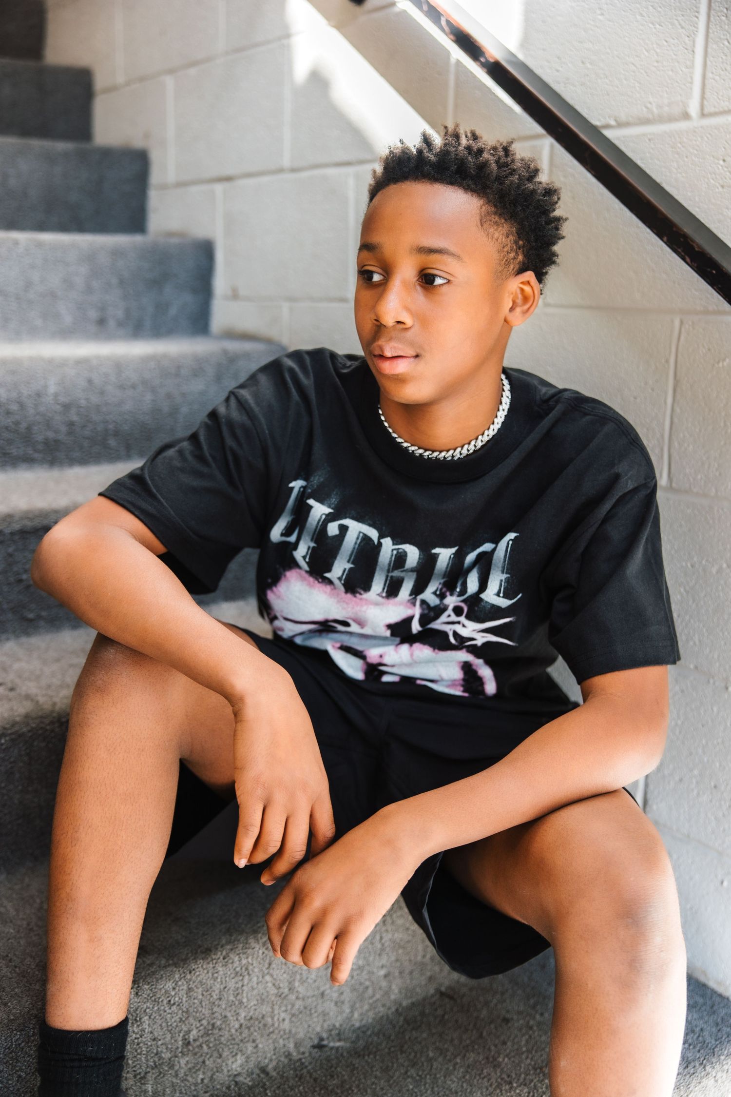 Young Black teenage boy in black graphic tee sitting on concrete stairs during urban portrait photography session.
