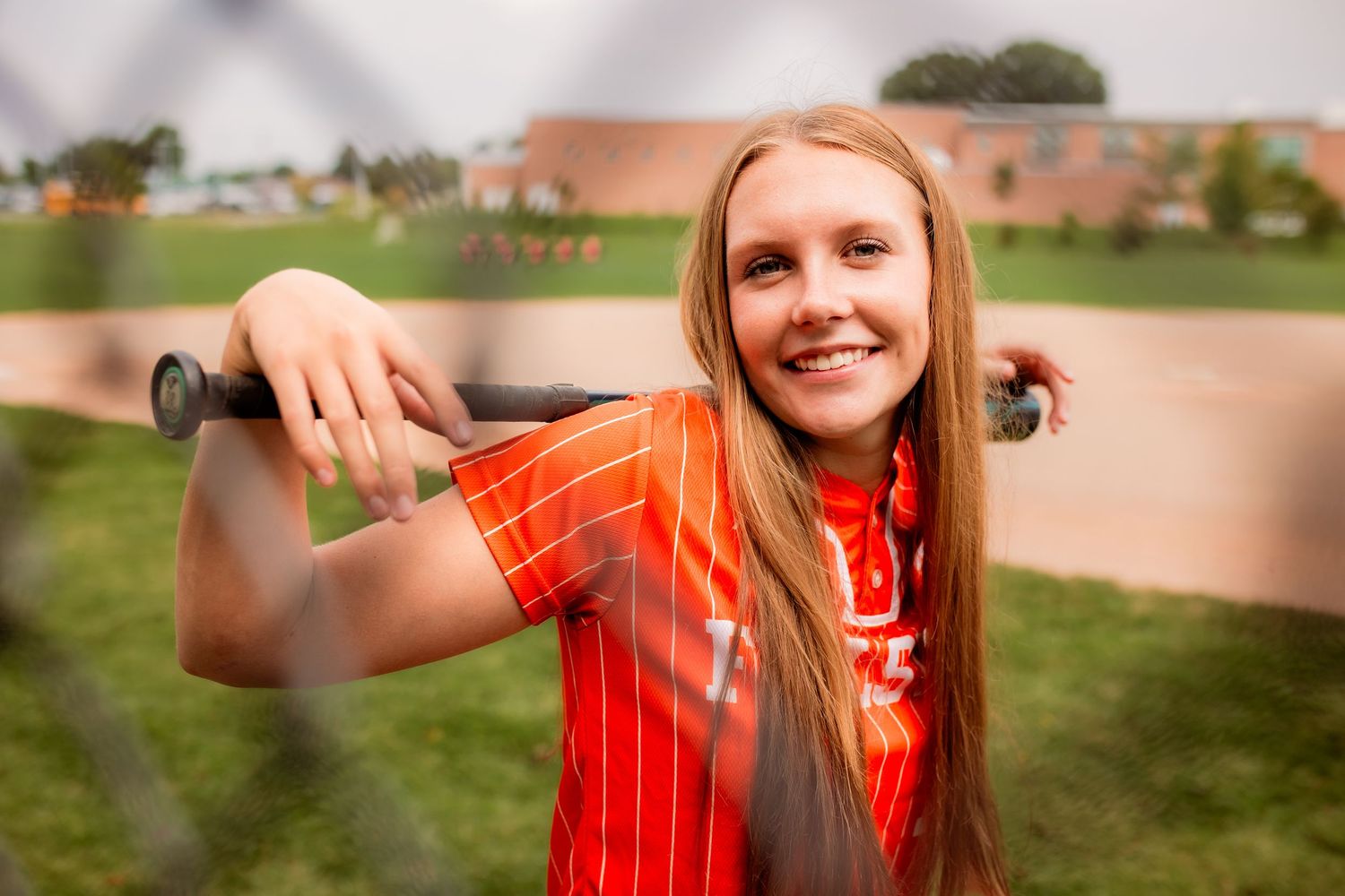 Smiling teen softball player in orange jersey leans on bat behind chain-link fence at baseball field.