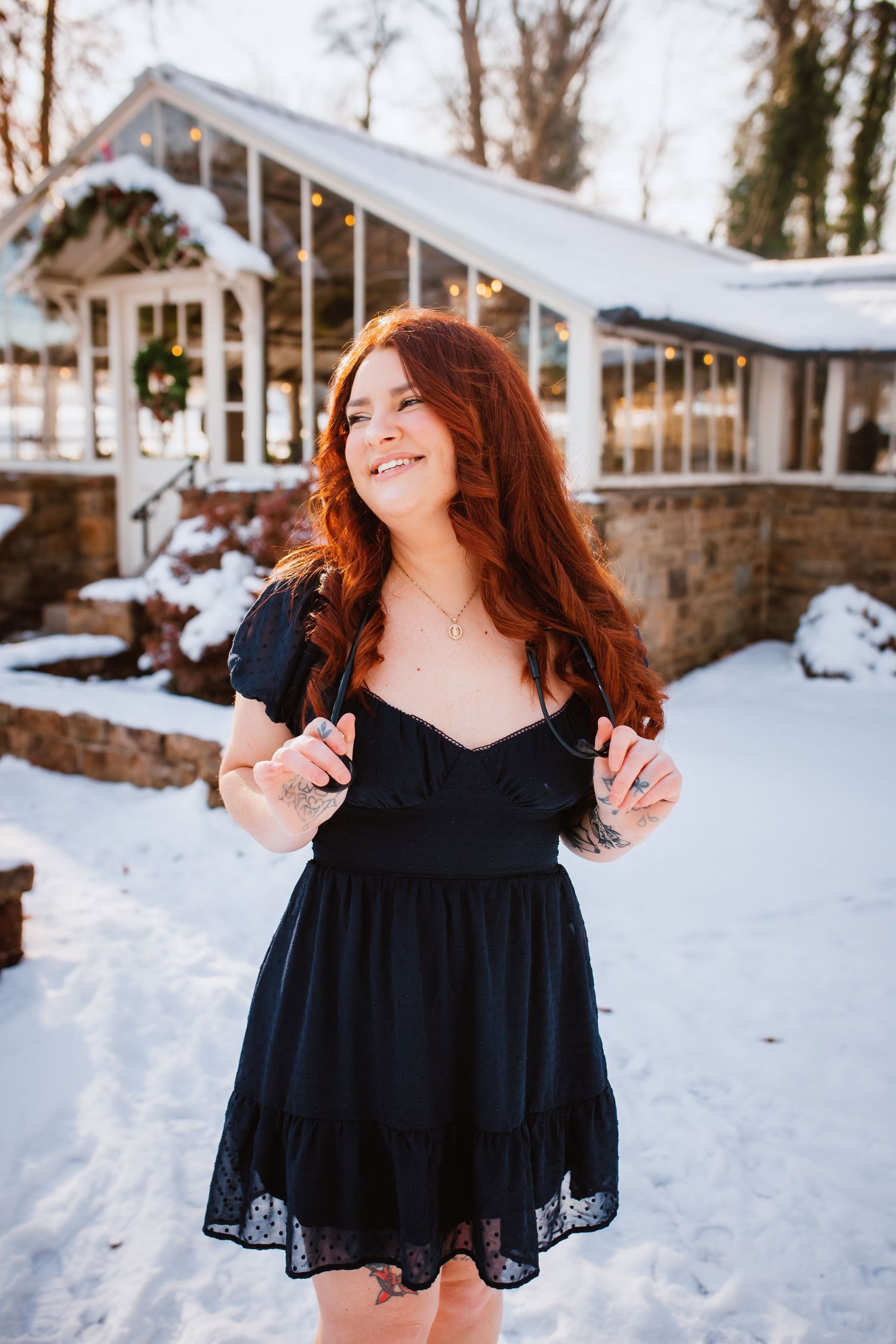 Smiling redhead woman in black dress stands in snow-covered garden with holiday decor.
