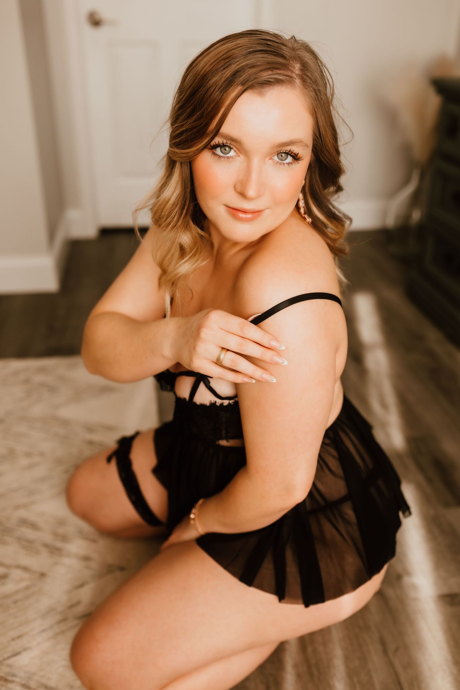 Blonde woman in black lingerie with garter belt poses on floor looking up.