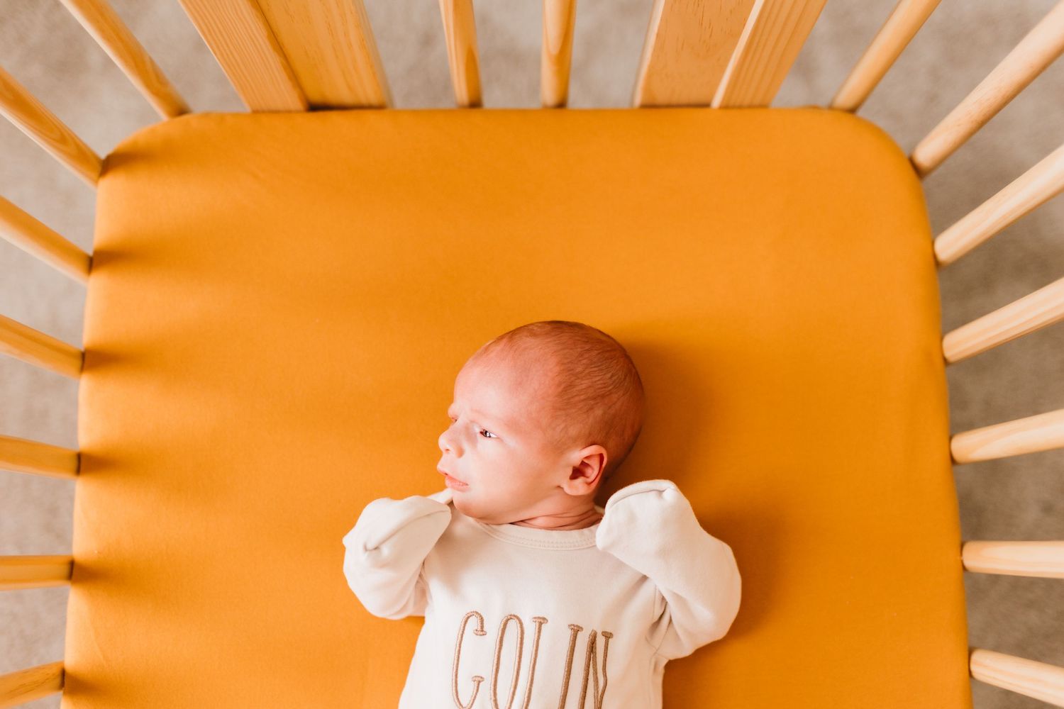 Newborn baby named Colin lying in an orange-sheeted wooden crib wearing a white onesie.