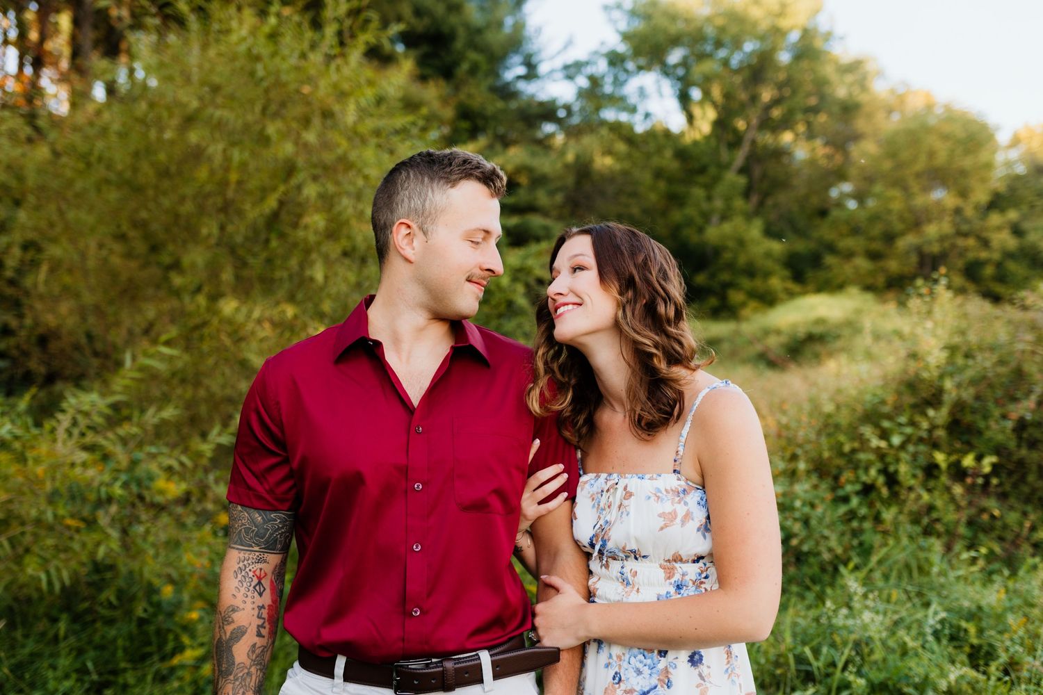 Couple gazing at each other smiling in a sunlit green outdoor setting.