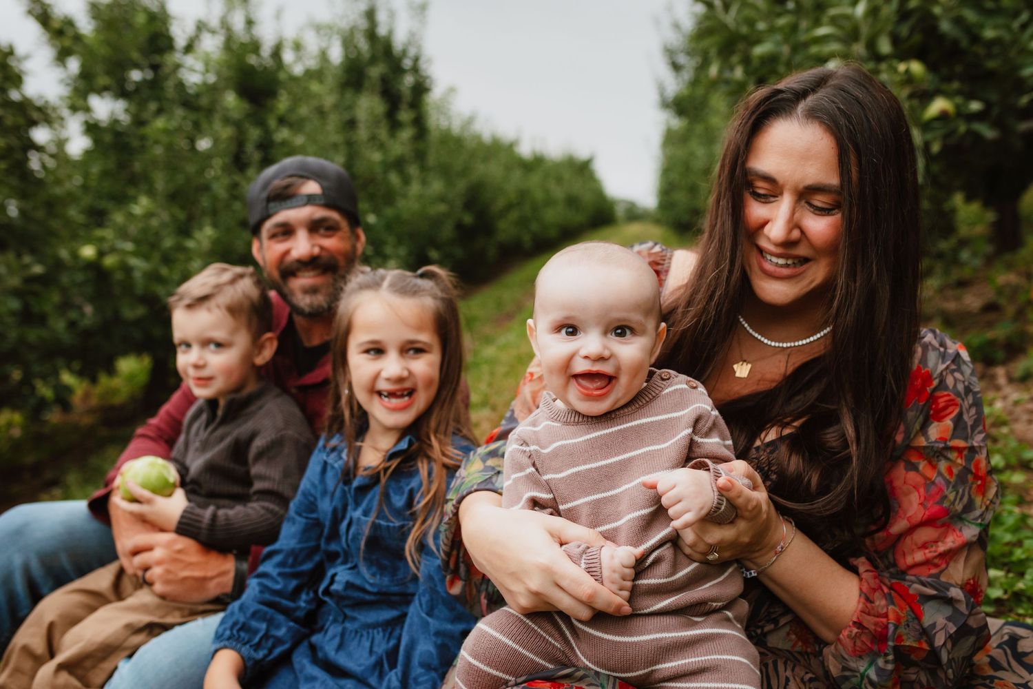 Smiling family of five sits together in an apple orchard, with a baby in a striped outfit, two young children, and two parents.