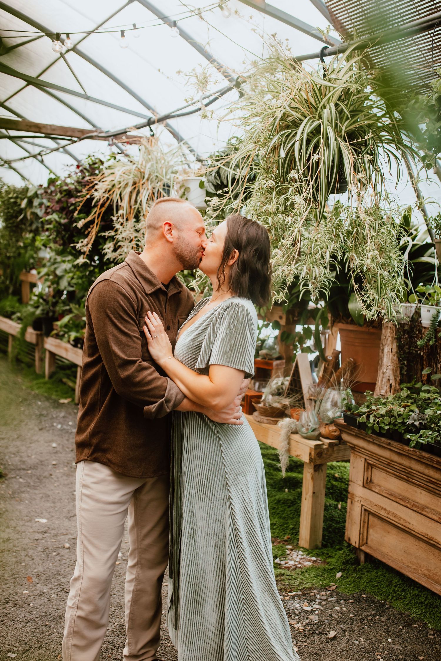 A couple shares a romantic kiss inside a lush greenhouse surrounded by hanging plants and potted greenery.