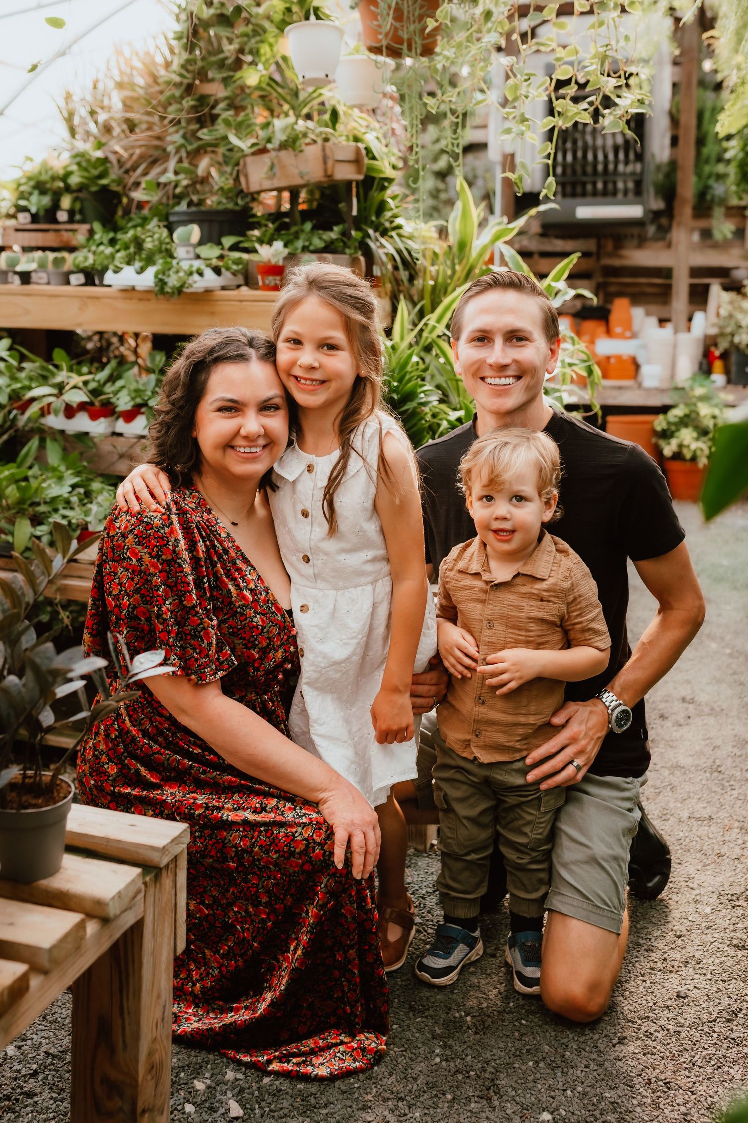 Happy family of four posing together in a lush greenhouse plant nursery, smiling for a portrait photo.