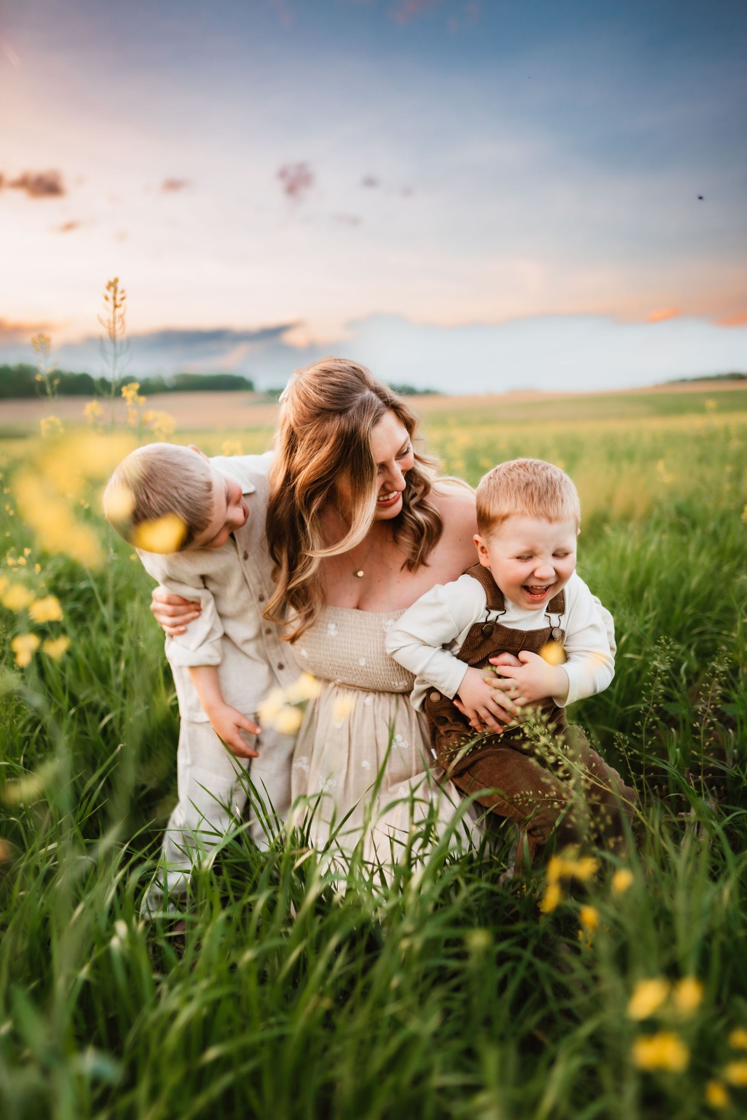Mother kneeling in green meadow at sunset, laughing with two young boys among yellow wildflowers.