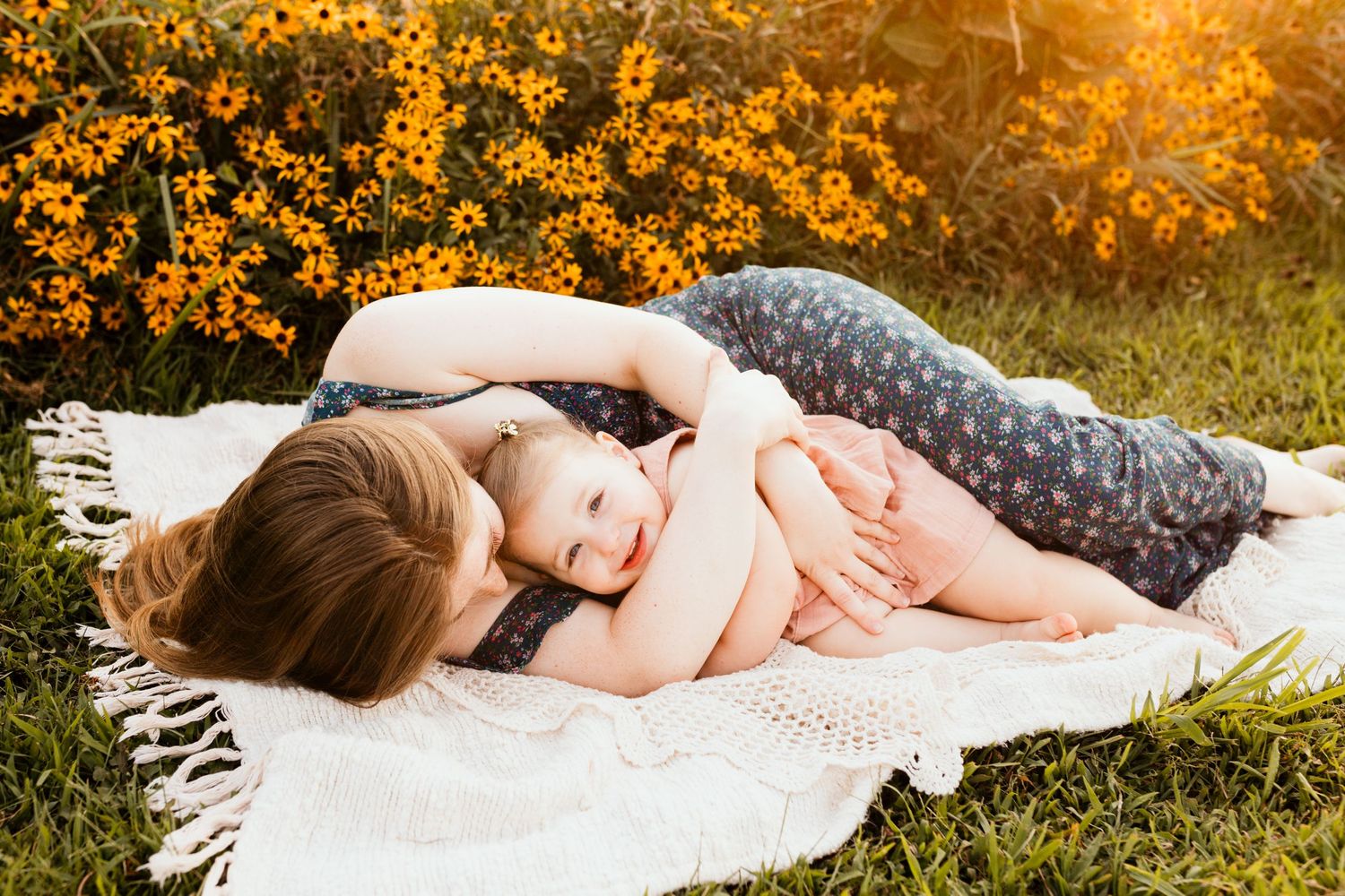 Mother lying on blanket outdoors hugging smiling baby near yellow flowers.