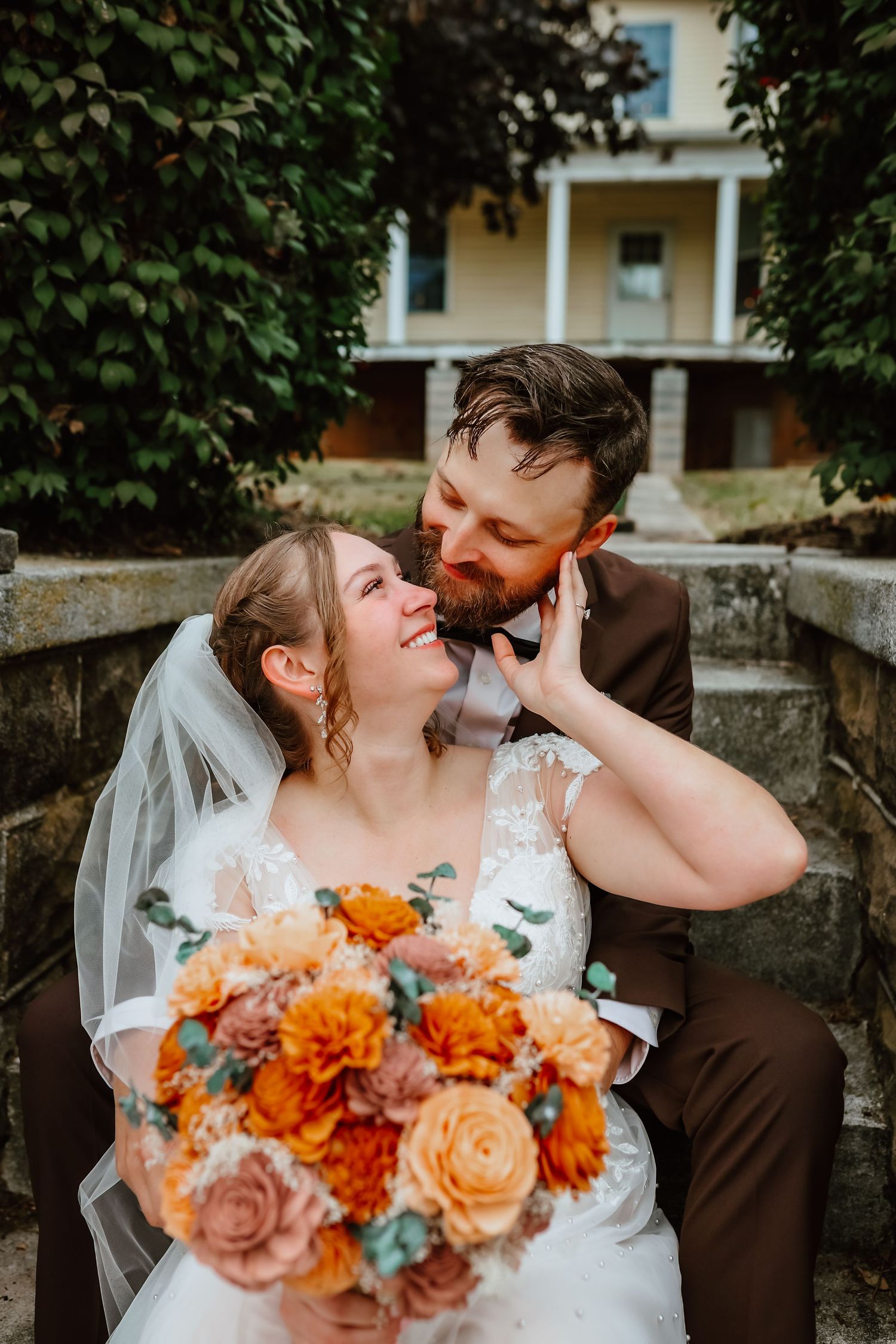 Bride holding orange bouquet smiling at bearded groom in brown suit outdoors.