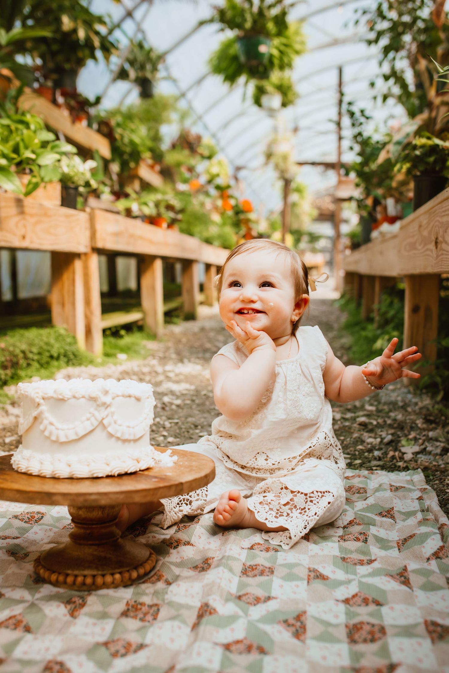 Smiling baby girl in white lace dress sitting beside a white cake on wooden stand in a greenhouse.