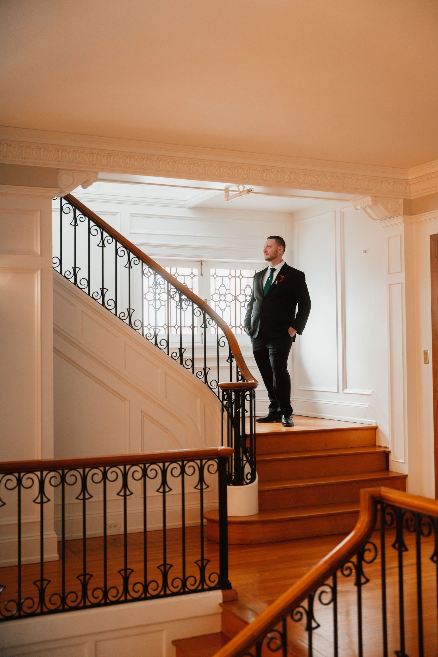 Groom in black suit stands on elegant curved staircase with wrought iron railings in a grand venue.