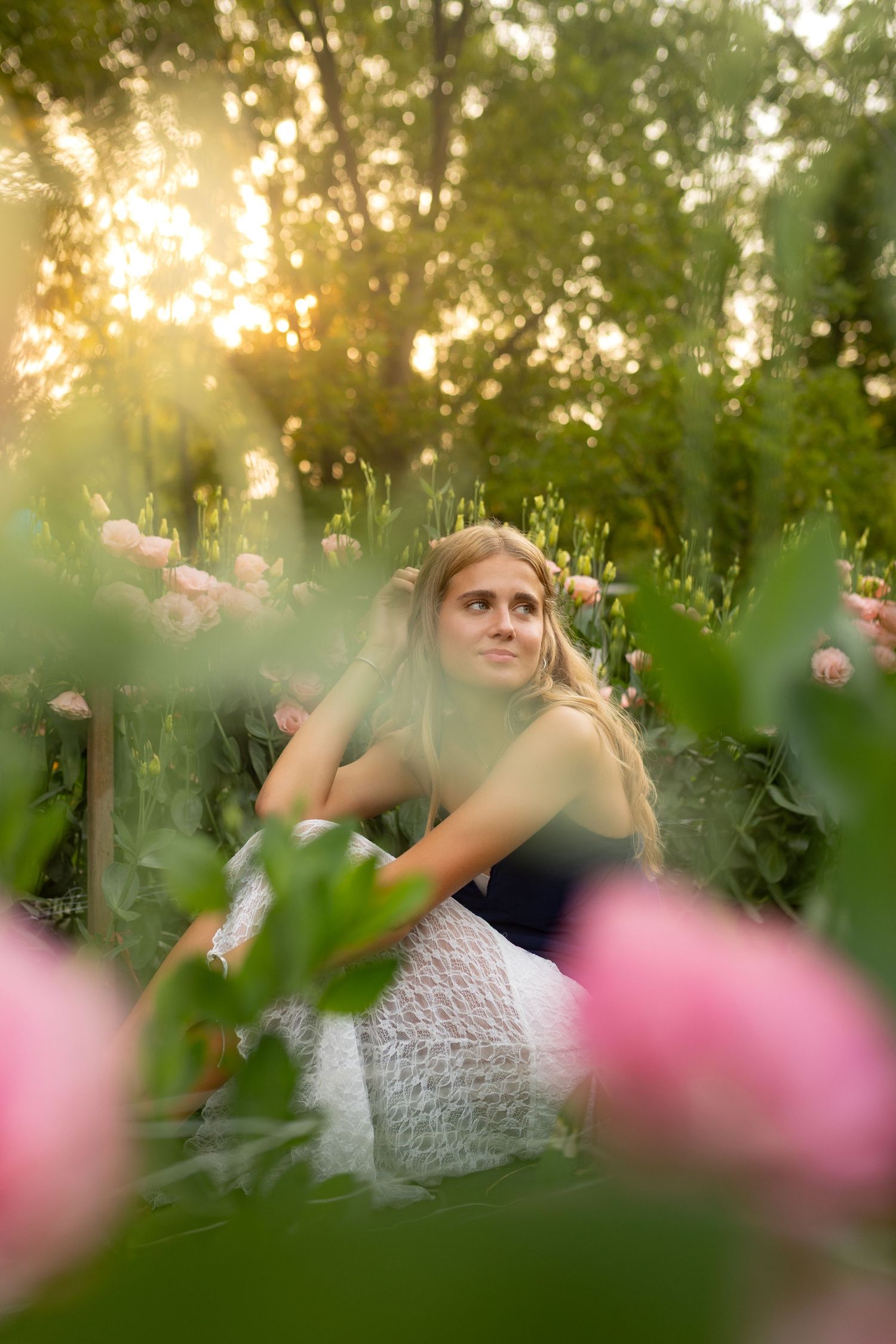 Young woman in white dress sitting among pink tulips in a sunlit garden at golden hour.