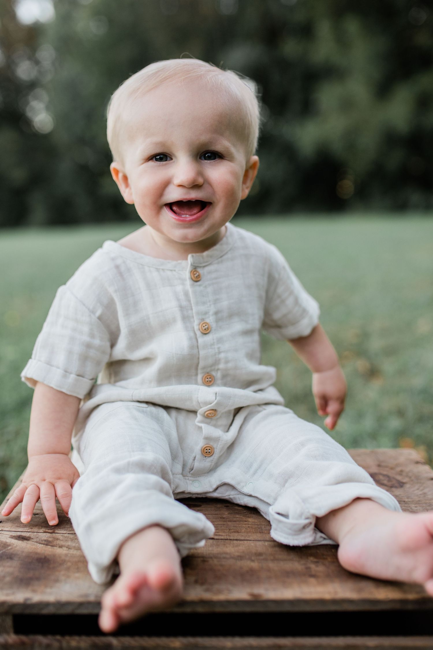 Happy smiling baby in neutral linen outfit sitting on wooden surface outdoors in green park setting.