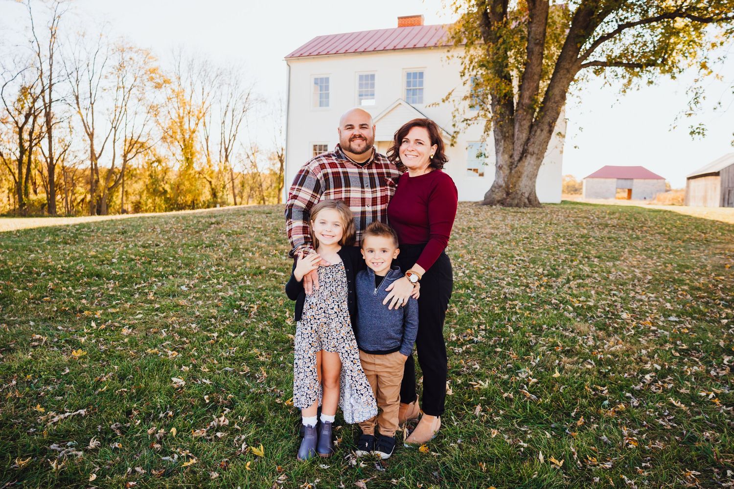 Family of four posing outdoors in fall foliage near a white historic building on a sunny autumn day.