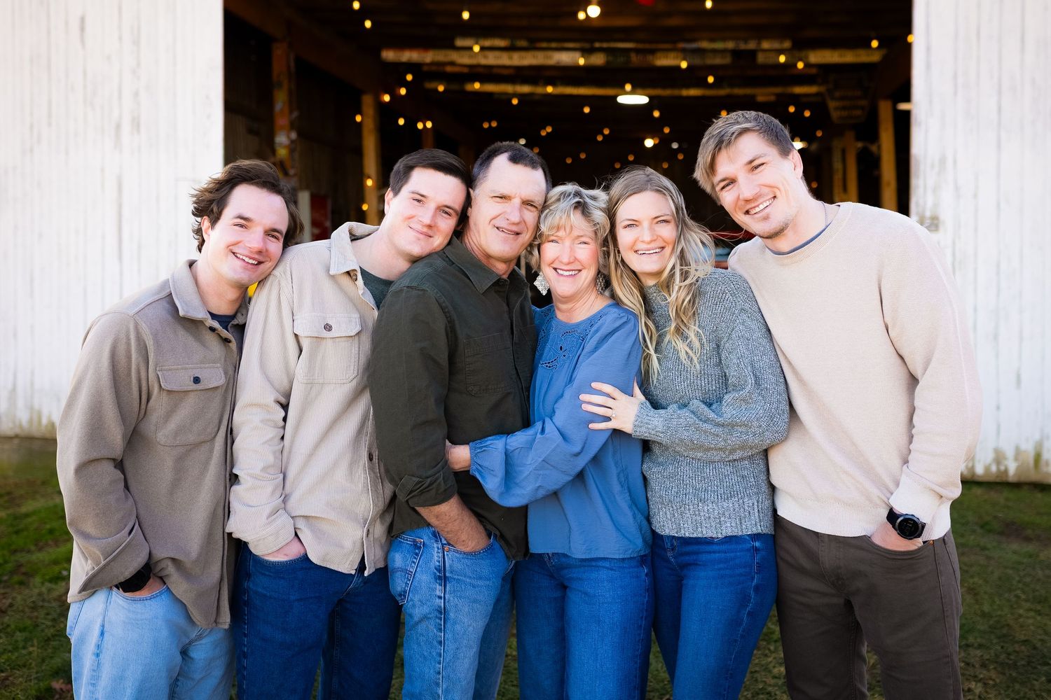 Family of six adults hugging and smiling together in front of rustic white barn.