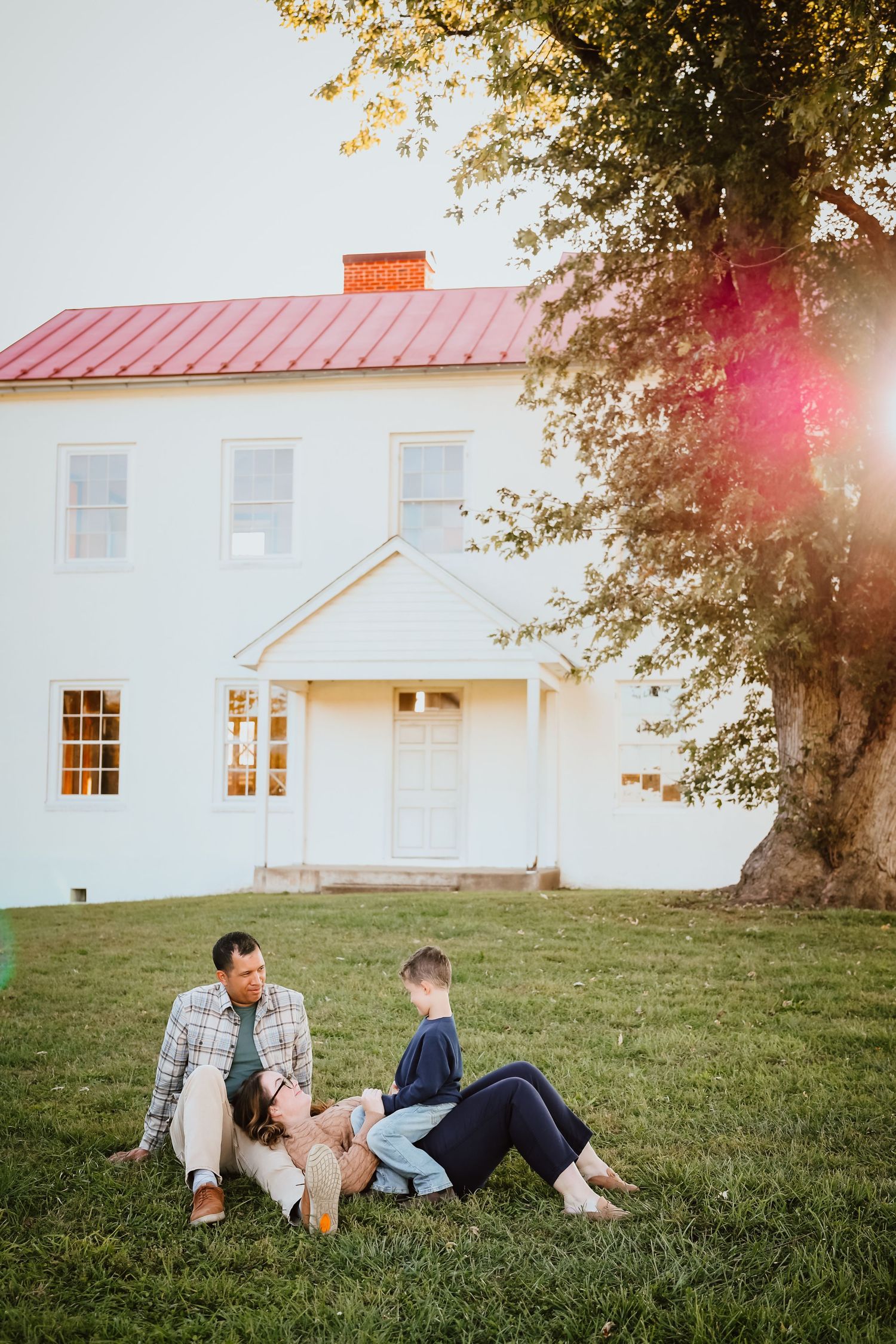 Family of three relaxing on green grass beside a white farmhouse with red roof.