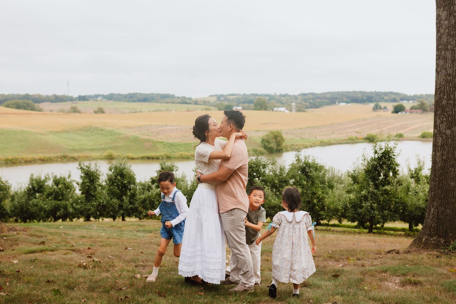 A couple embraces in a grassy orchard while three young children stand beside them, with a pond and rolling countryside in the background.