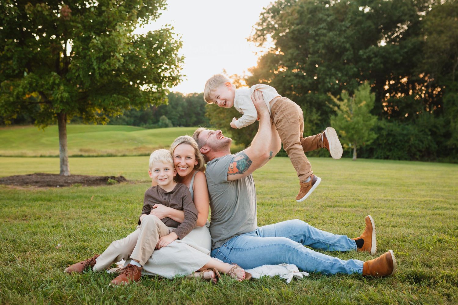 Family of four enjoying a sunny outdoor photo session on green grass, parents playing with two young children at golden hour.