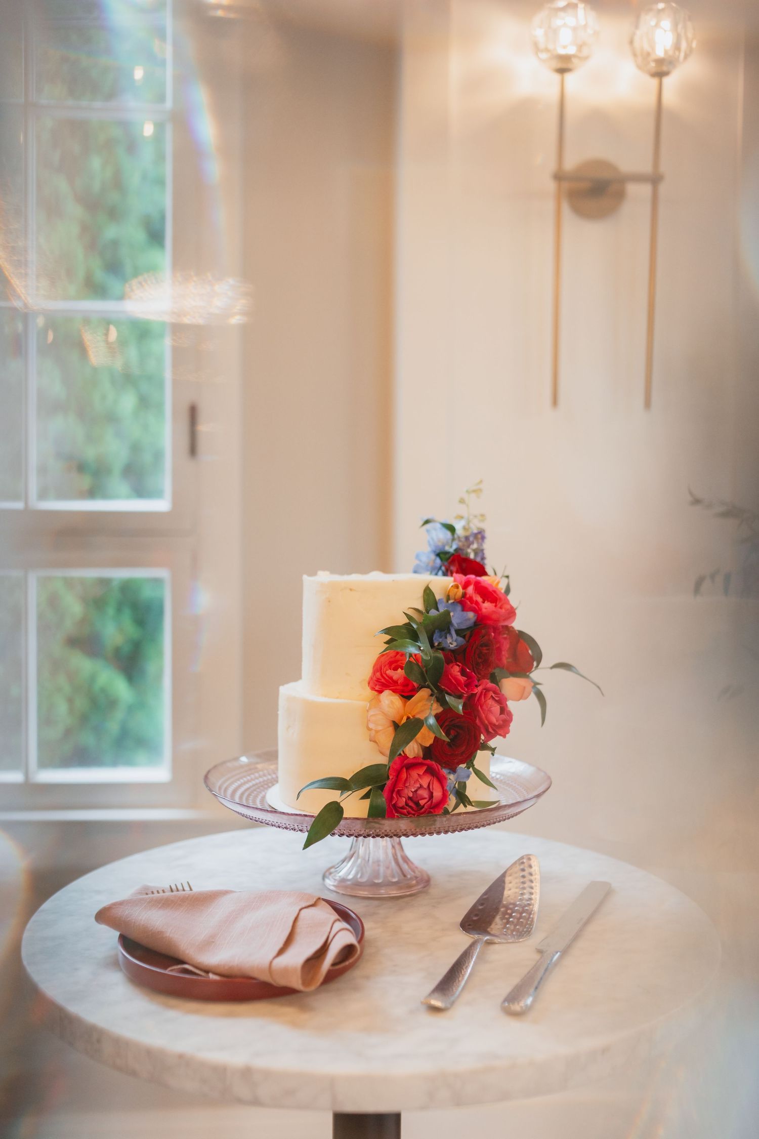 Two-tier white wedding cake adorned with red peonies and greenery on marble table with serving utensils.