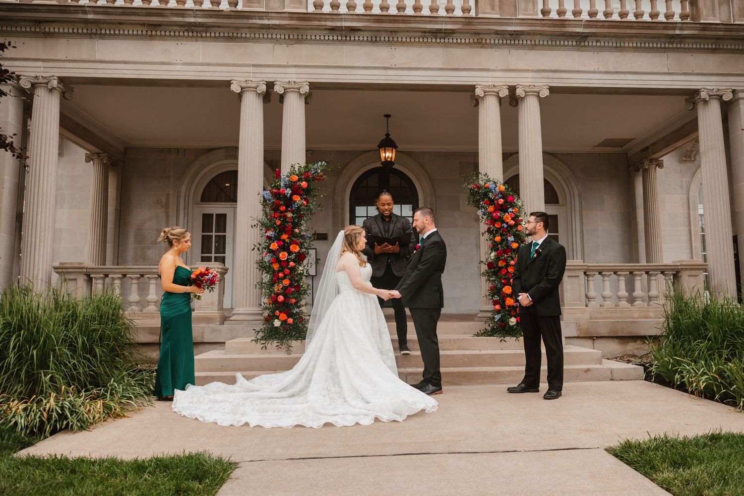 Bride and groom exchange vows outdoors before a grand columned mansion with vibrant floral arrangements.