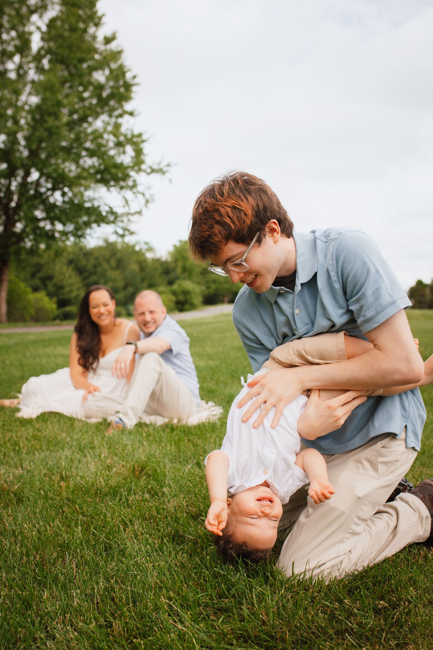 Father playfully holding baby upside down on grass while mother and toddler watch and smile in park.