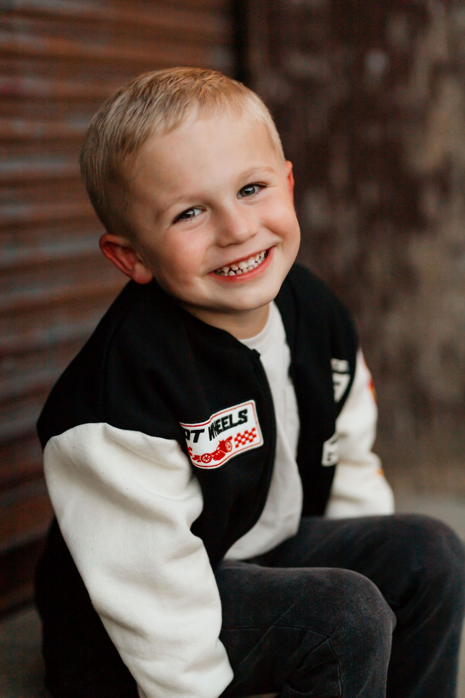 Cheerful young boy smiling in black and white Hot Wheels jacket posing against rustic brick background.