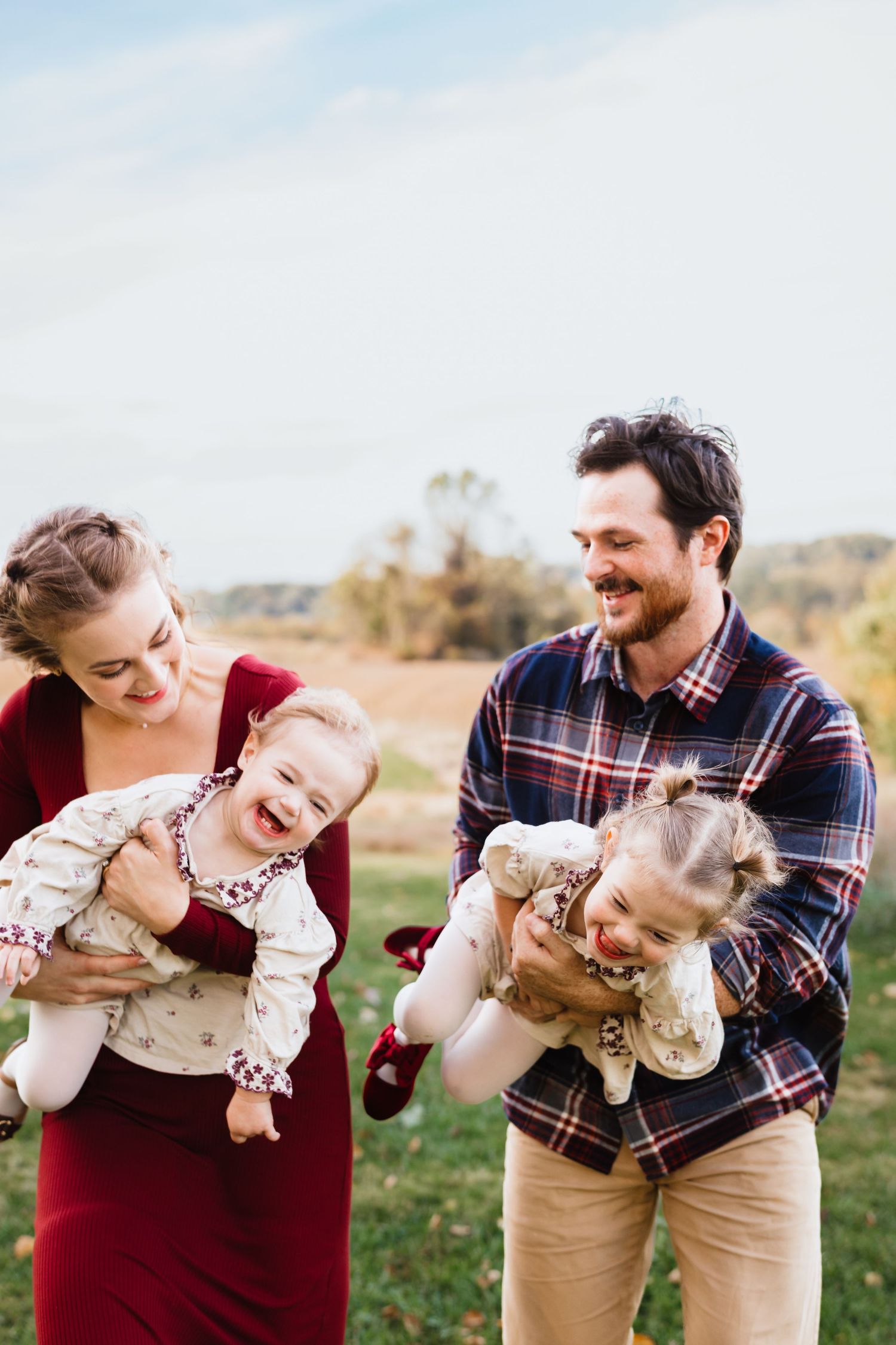 Young family laughing outdoors in autumn field, parents holding two toddlers in floral outfits during fall family photo session.
