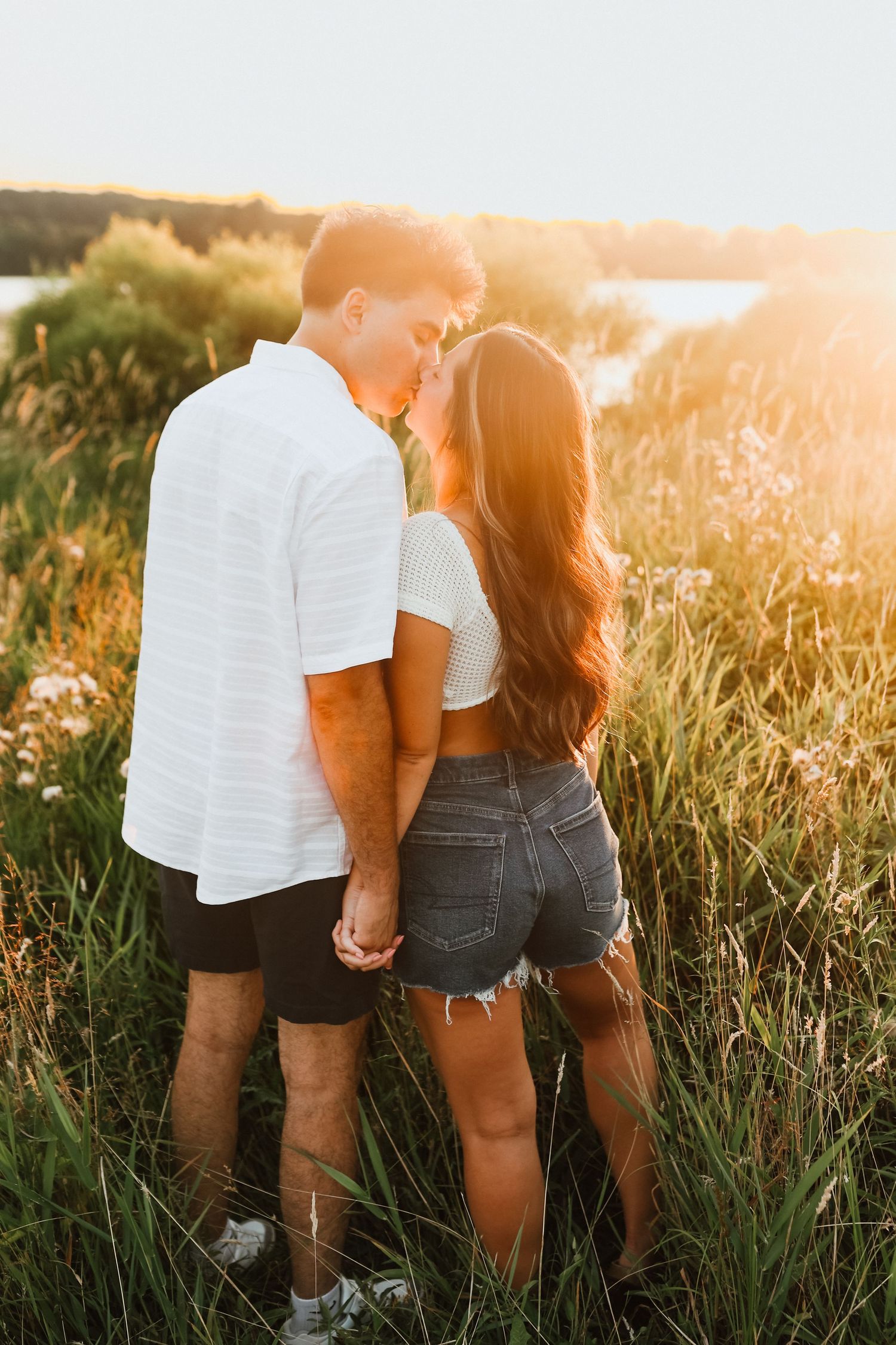Couple holding hands in golden sunset light among tall grass near a lake.