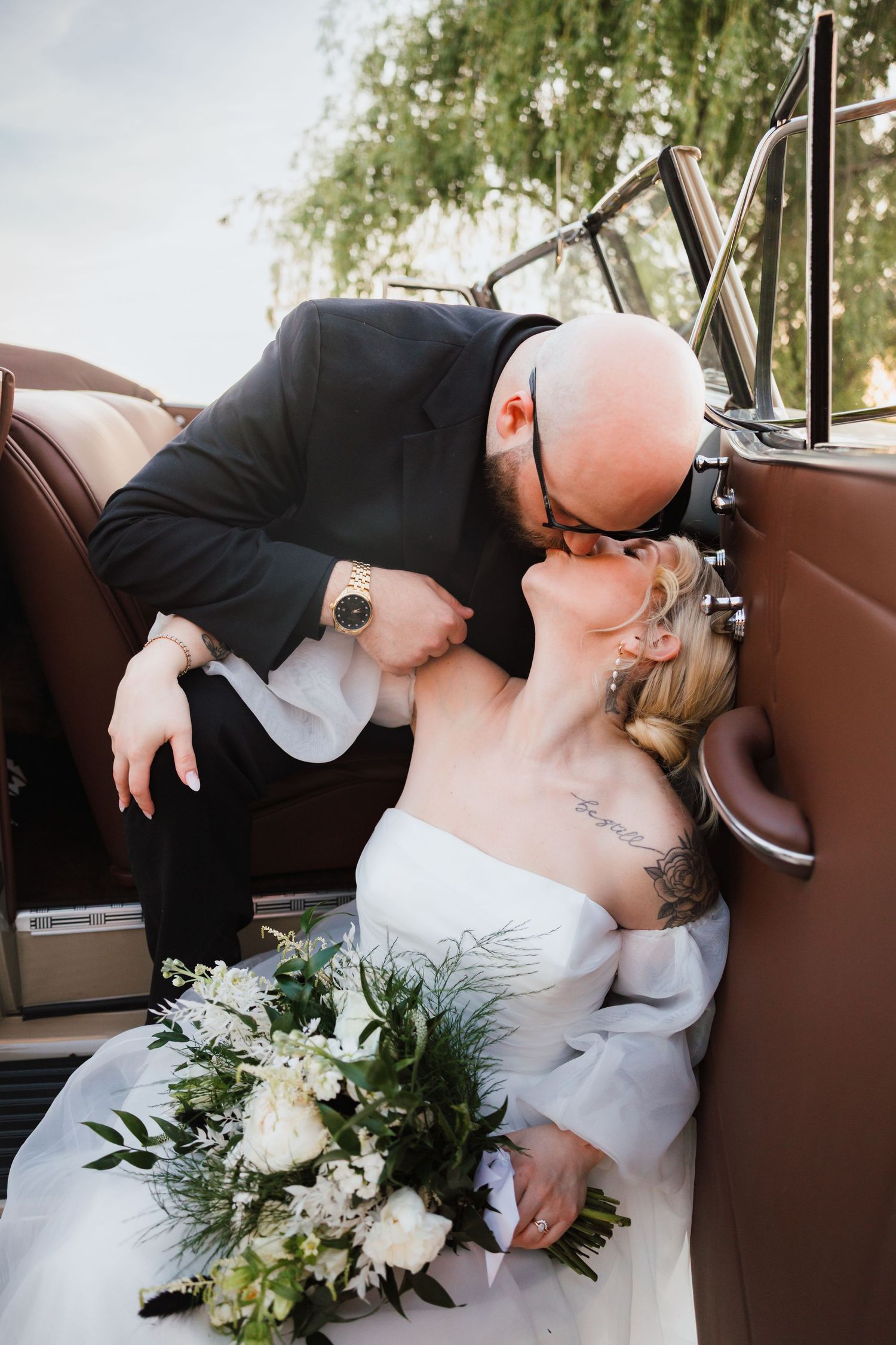 Bride and groom share a romantic kiss inside a vintage car, bride holding a white and green bouquet.