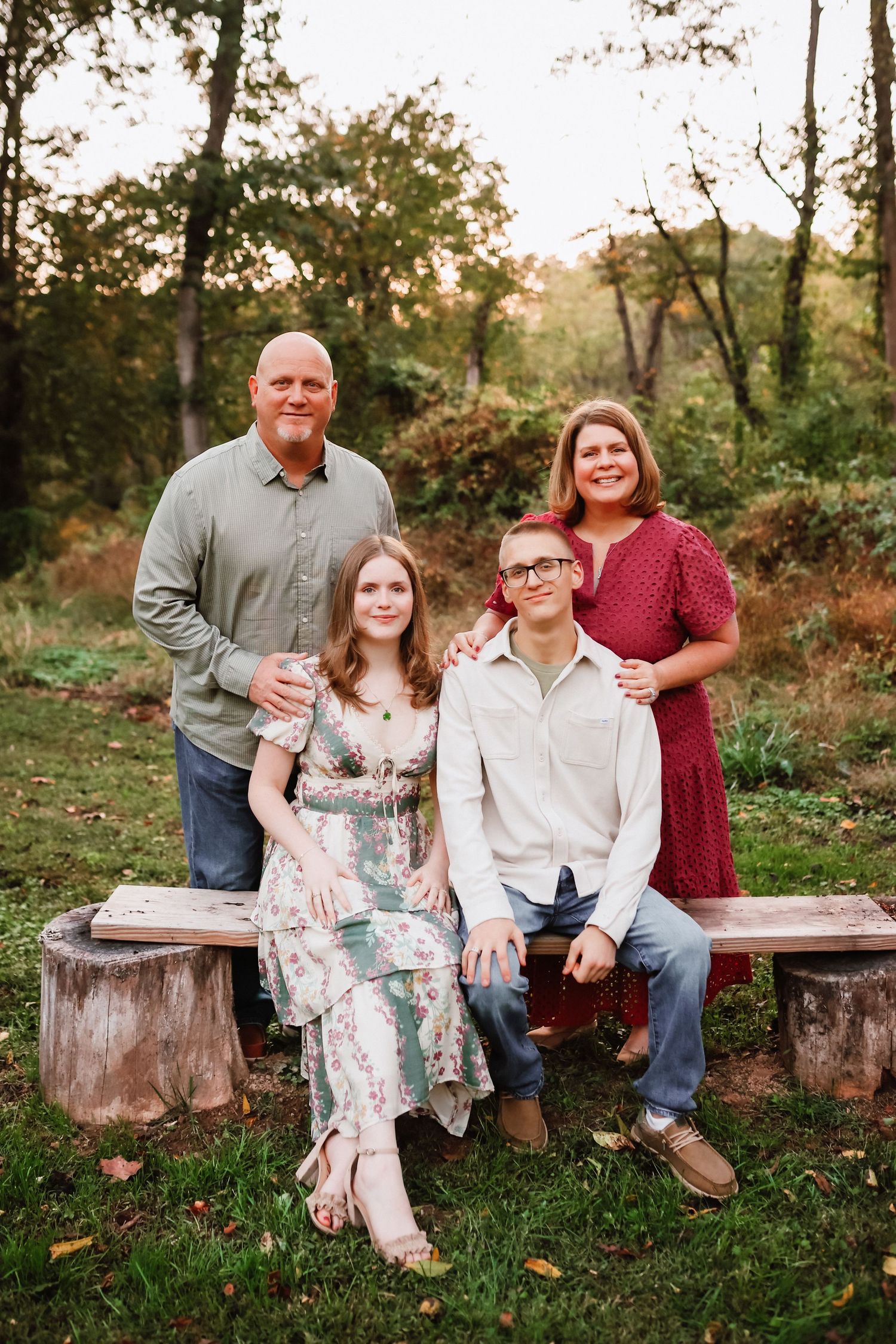 Family of four poses on a rustic wooden bench surrounded by autumn woodland scenery.