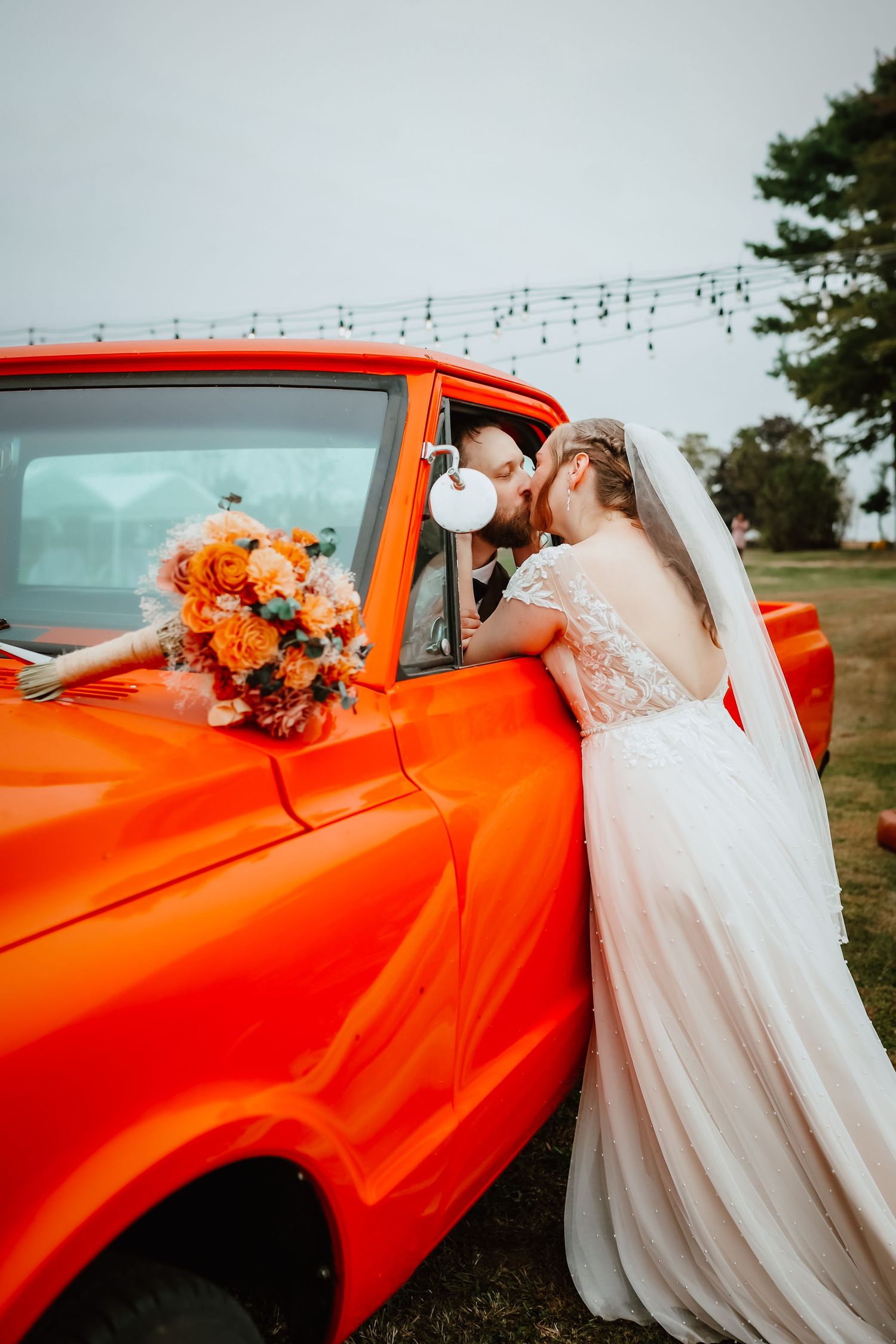 Bride and groom sharing a kiss through the window of a bright orange vintage pickup truck.