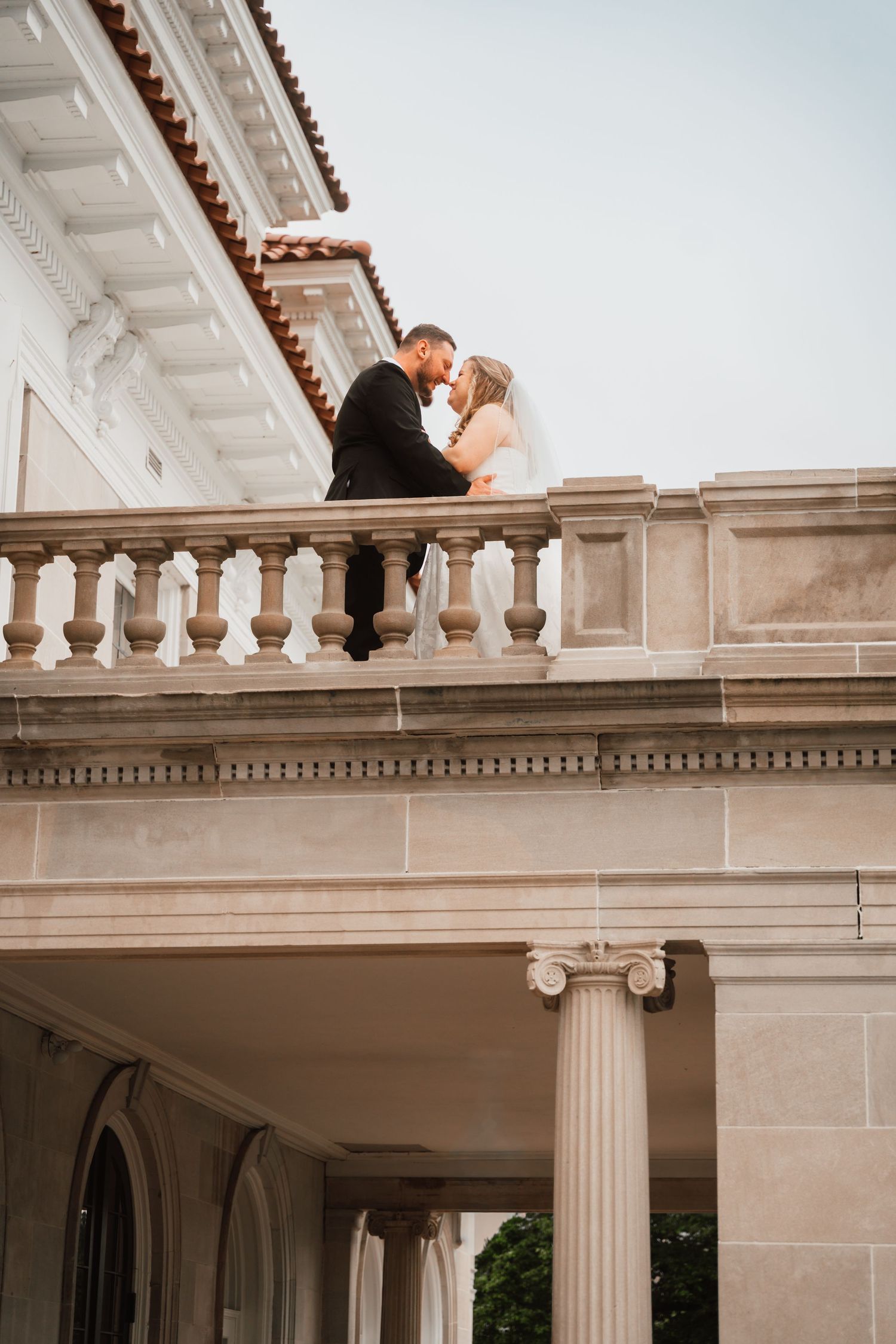 Bride and groom share romantic moment on ornate stone balcony of a classical wedding venue.