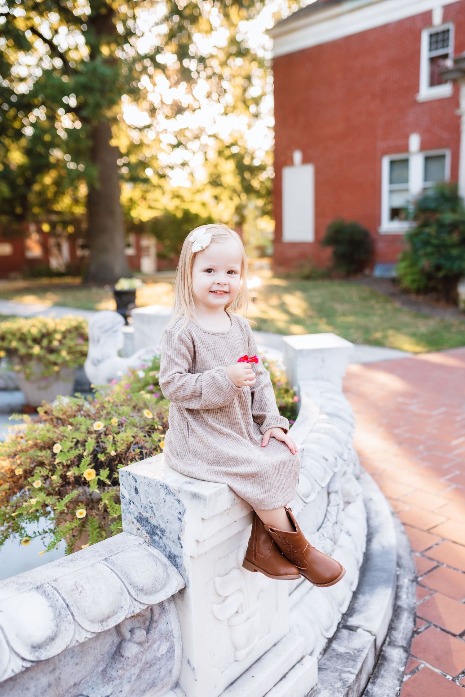 Adorable blonde toddler girl in beige dress and boots sitting on stone garden fountain.