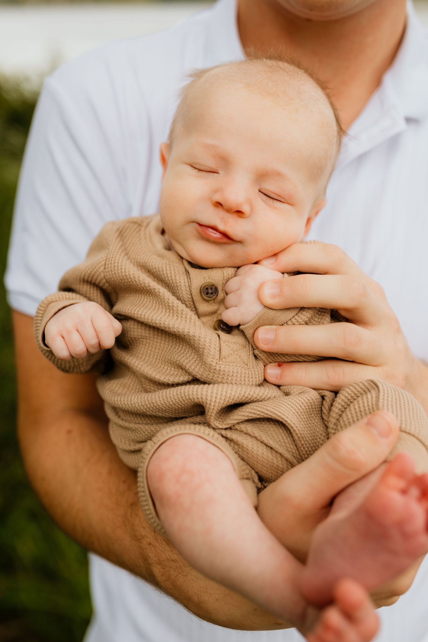 Peaceful newborn baby sleeping in fathers arms wearing tan knit outfit outdoors.