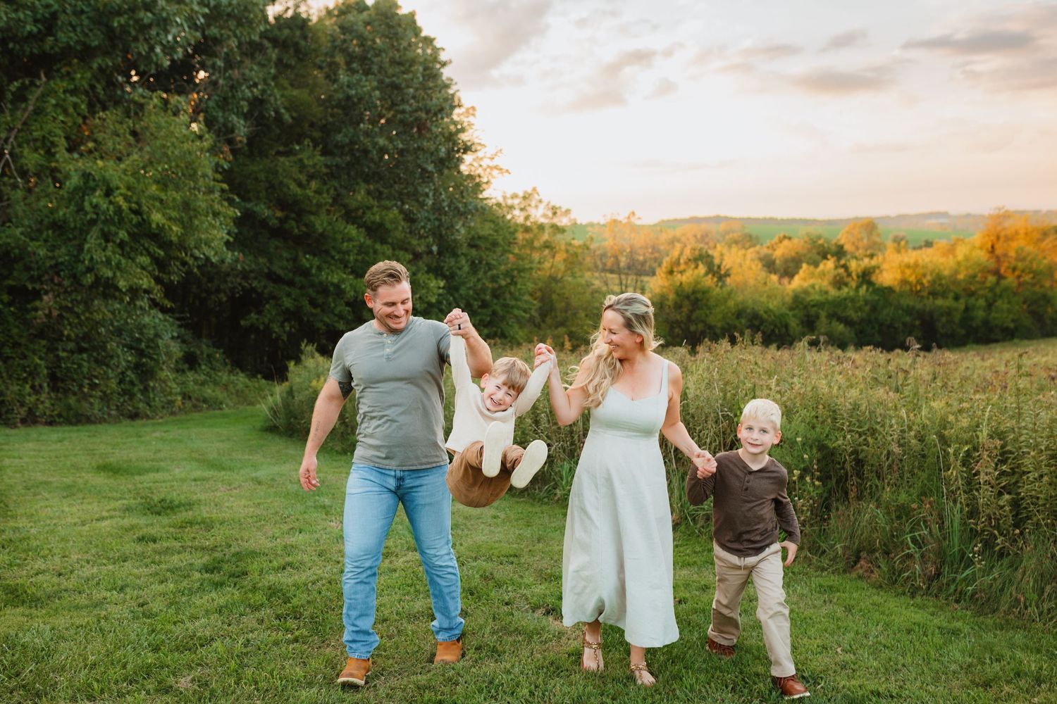 Family of four playing outdoors at sunset, parents swinging child between them on lush green field.